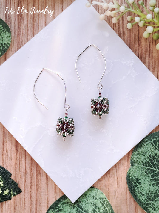 Pair of red and green cube beaded earrings on a white tile with green leaves and flowers in the background.