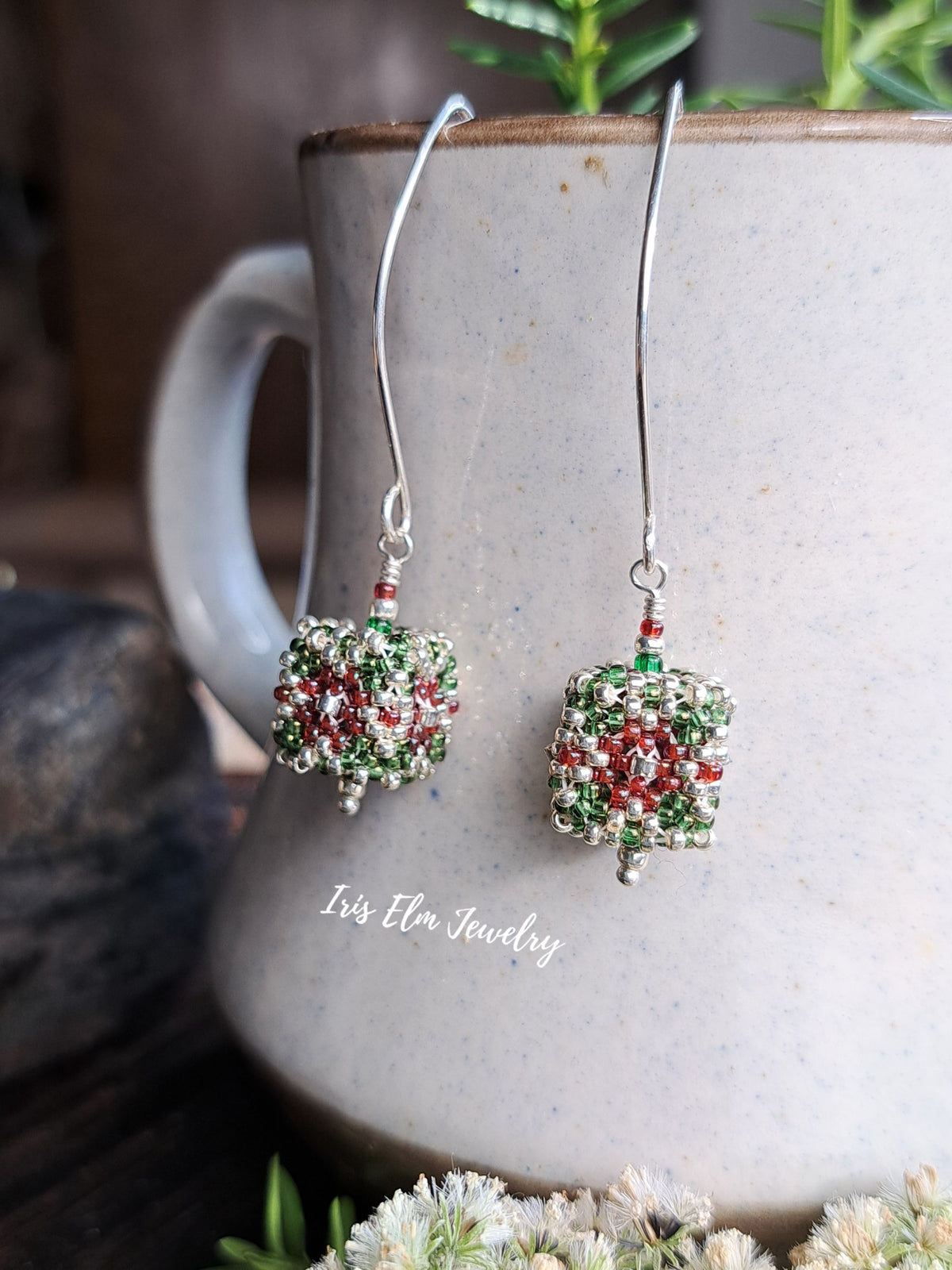 A pair of handmade beaded dangle earrings with silver hooks and green, red, and silver seed bead patterns, hanging from a speckled gray ceramic mug. The text 'Iris Elm Jewelry' is printed on the front of the mug.