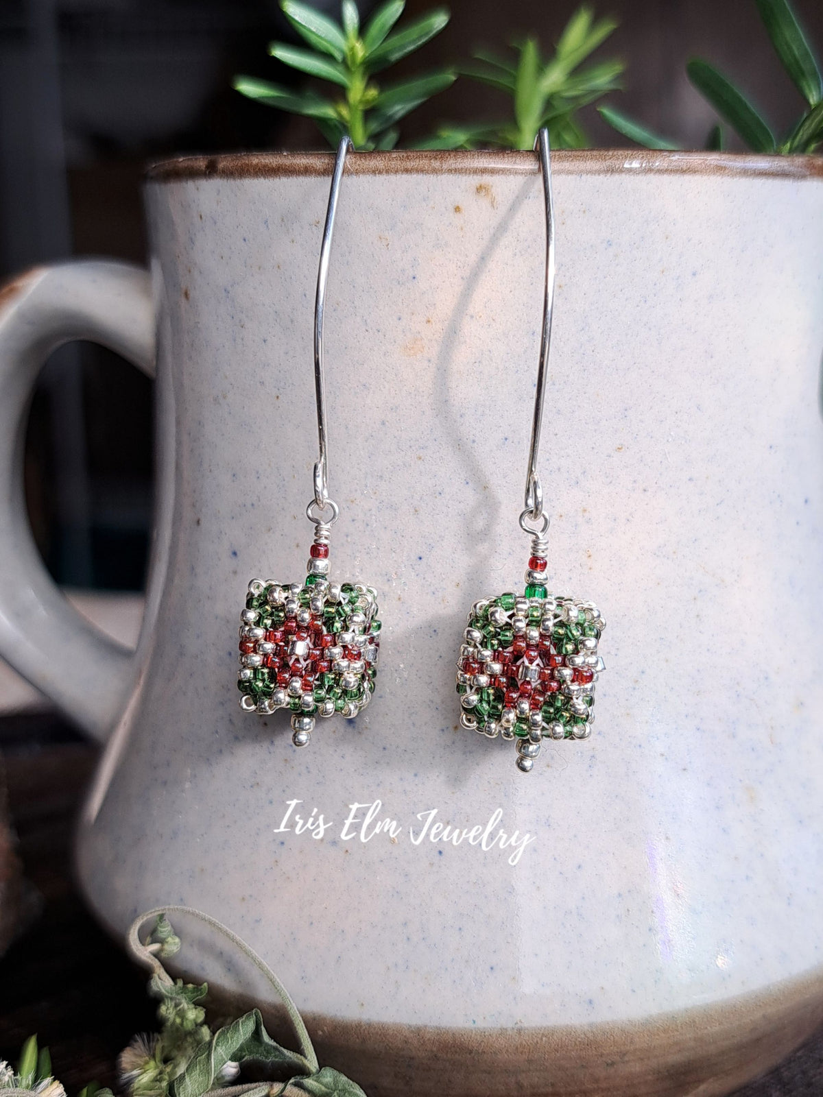 A pair of handmade beaded dangle earrings with silver fishhook wires and square charms decorated with red, green, and silver seed beads, hanging in front of a ceramic mug with green plants in the background. The text 'Iris Elm Jewelry' is visible on the mug.