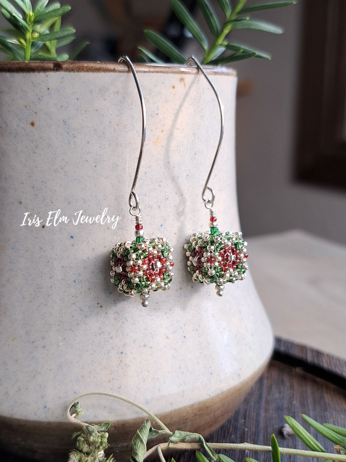 Handmade beaded dangle earrings with silver fishhook wires, featuring red, green, and silver seed beads in a holiday ornament design, displayed in front of a ceramic pot.
