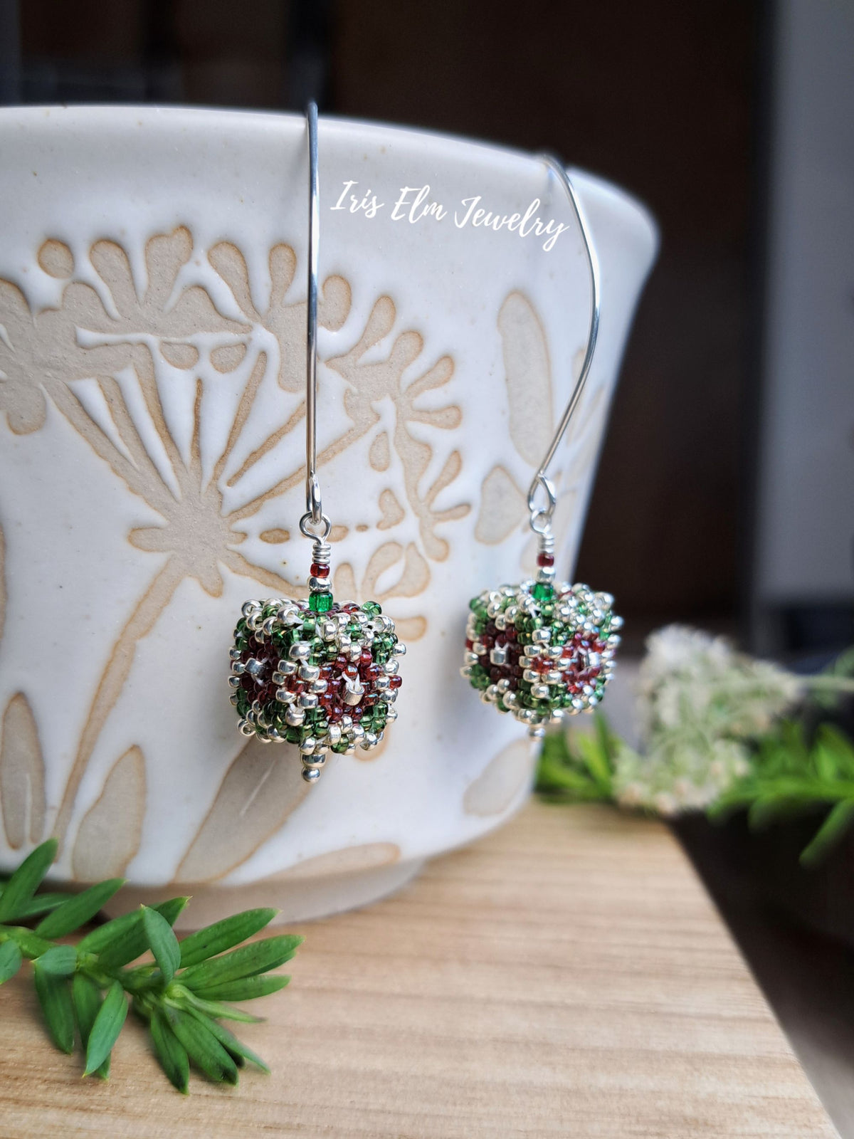 A pair of beaded drop earrings with green and burgundy seed beads, suspended from silver fishhook wires, displayed in front of a white ceramic mug on a wooden surface.