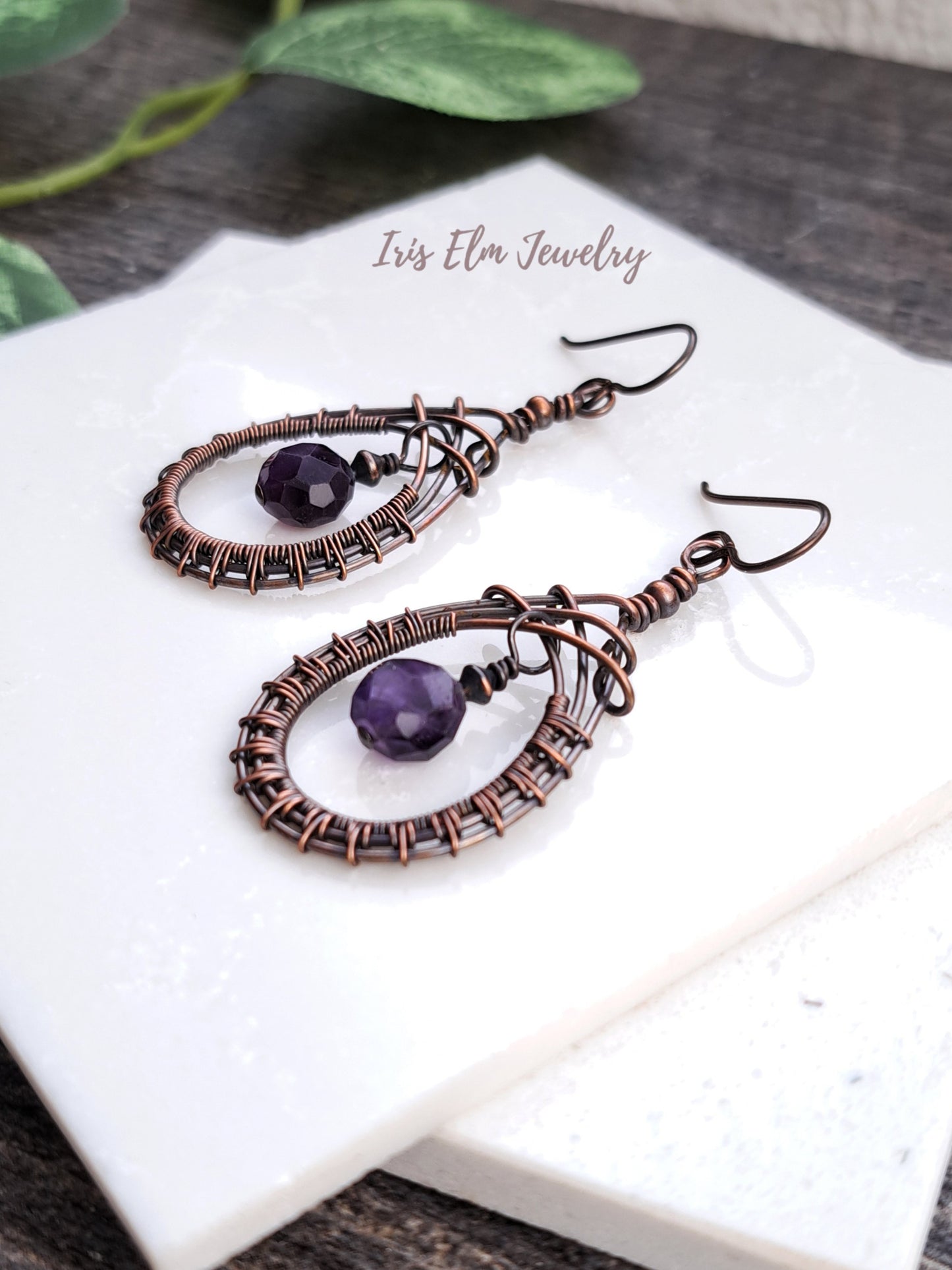 Pair of wire-wrapped teardrop earrings with purple amethyst stones on a white surface, branded 'Iris Elm Jewelry'.