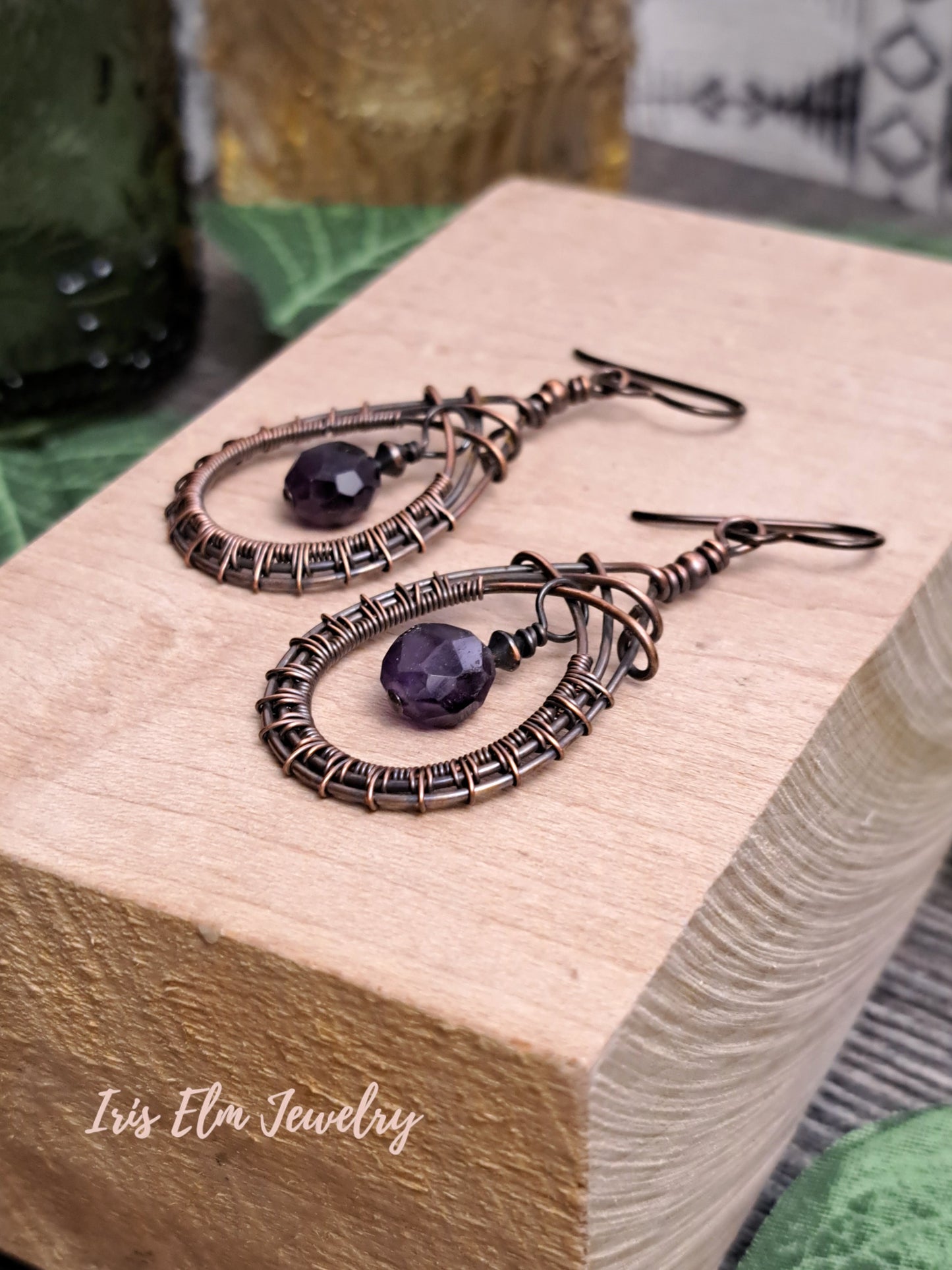 Pair of copper wire-wrapped teardrop earrings with faceted amethyst purple stones on a wooden block, branded 'Iris Elin Jewelry'.