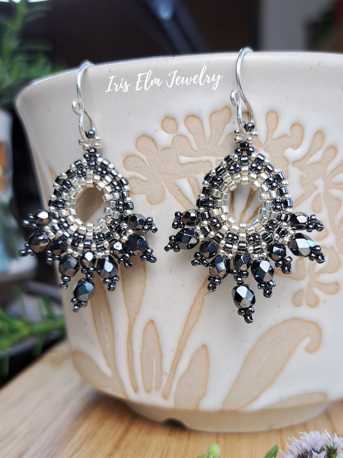 A pair of dangling earrings with silver-toned and dark gray faceted beads, displayed on a beige ceramic cup. The brand name 'Iris Elm Jewelry' is printed in white script across the top.