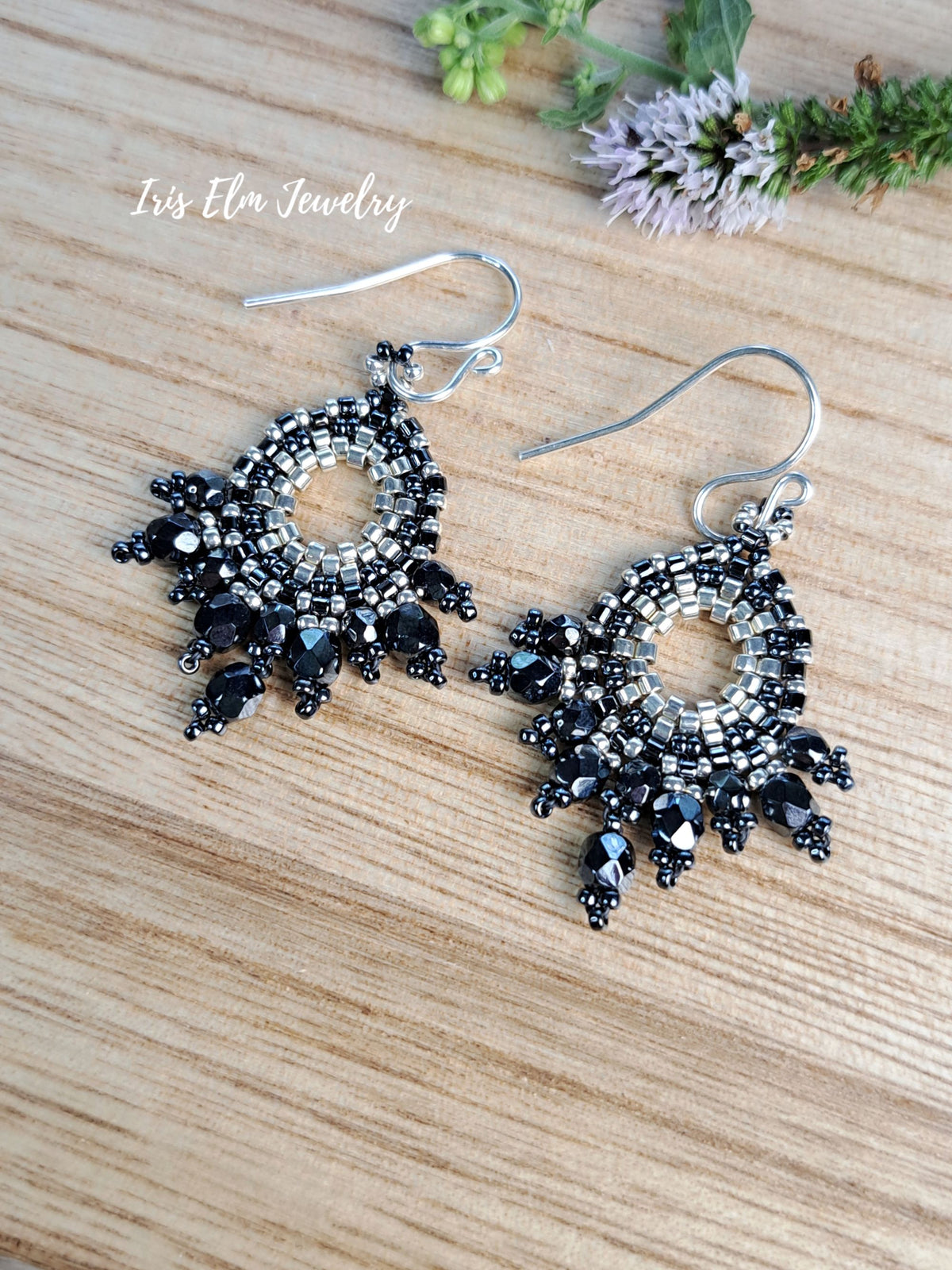A pair of bohemian-style beaded dangle earrings with black and silver beads, silver fishhook wires, and dangling black teardrop beads, displayed on a light wood grain surface with small flowers in the corner. The text 'Iris Elm Jewelry' is visible in the upper left.
