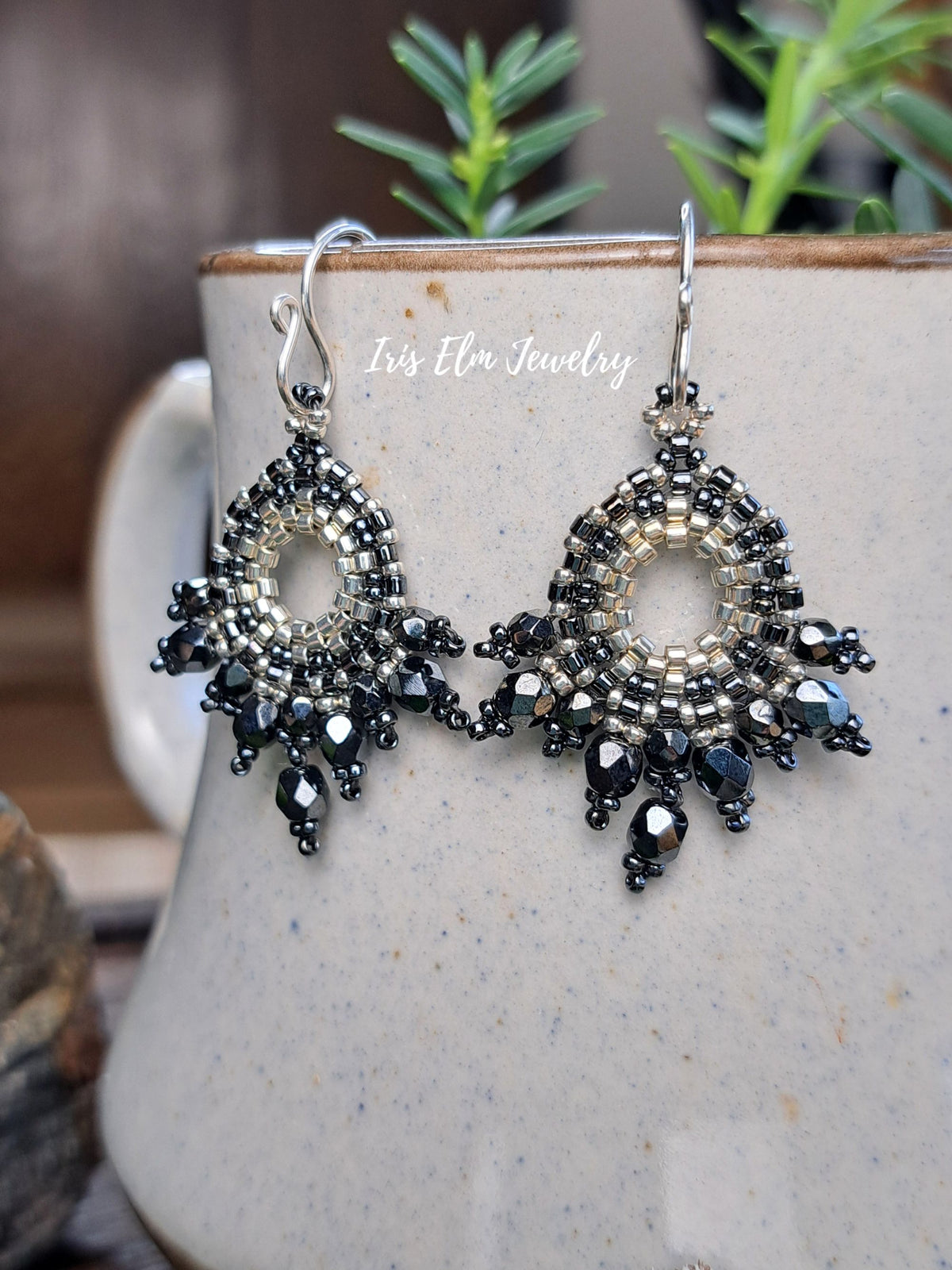A pair of intricate beaded dangle earrings with pewter czech glass faceted beads and silver-toned metal hooks, displayed on a speckled ceramic mug with green plants in the background. The watermark 'Iris Elm Jewelry' is visible on the mug.