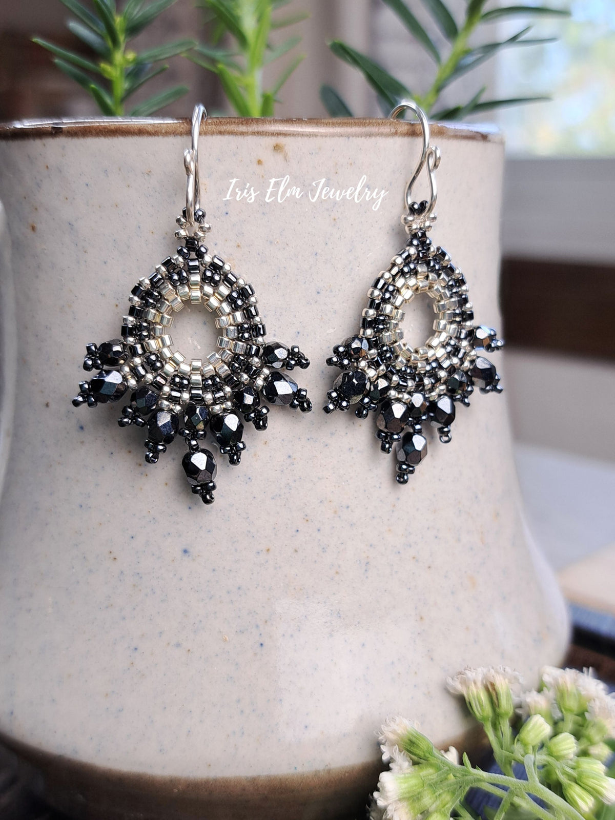 A pair of intricate beaded dangle earrings with black and silver beads, hanging from silver hooks on a speckled beige ceramic pot with green plants in the background.