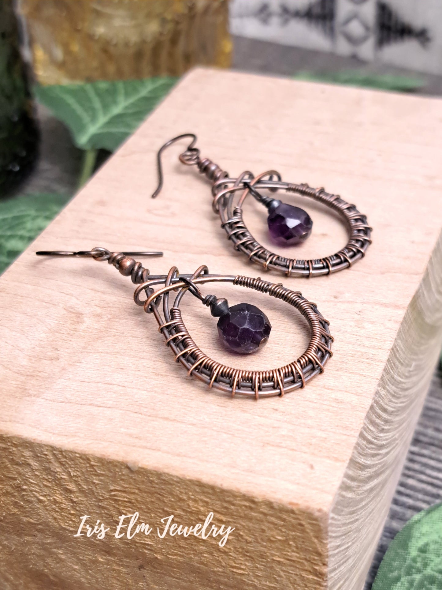 Pair of copper wire-wrapped earrings with purple amethyst beads on a wooden block, branded 'Iris Elm Jewelry'.