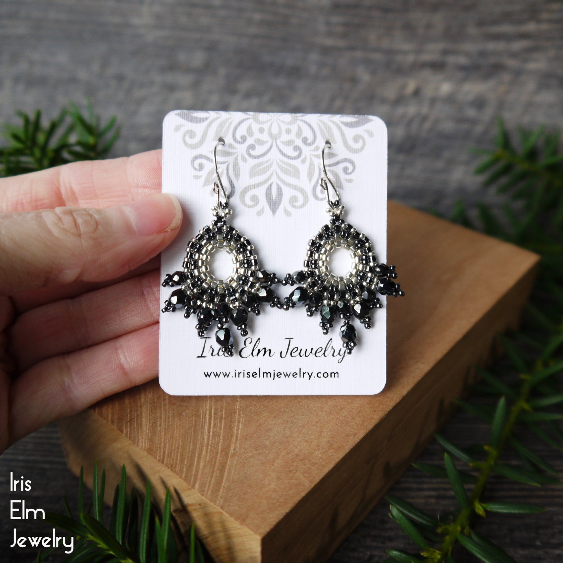 Pair of dark grey Czech glass handmade beaded earrings on a 'Iris Elm Jewelry' card held by a hand with a natural background.