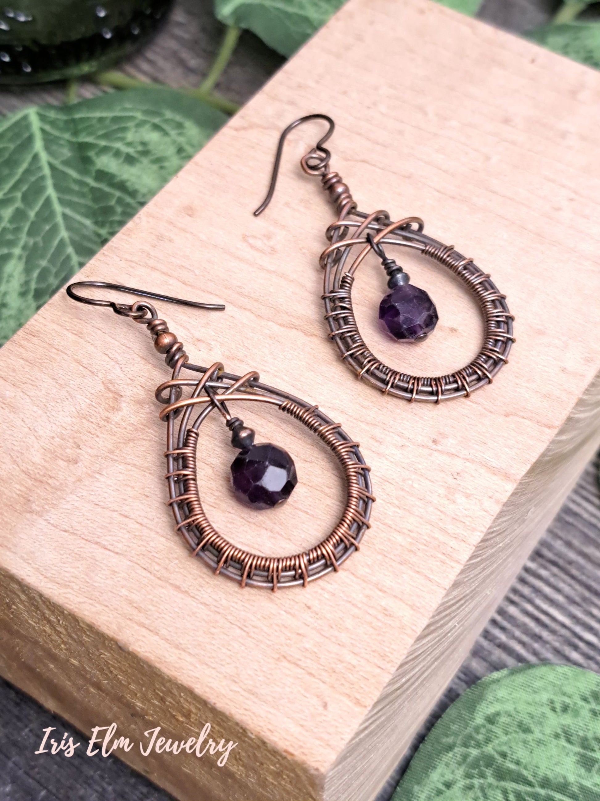 Pair of copper wire-wrapped earrings with dark purple amethyst beads on a wooden block, brand 'Iris Elm Jewelry' visible.