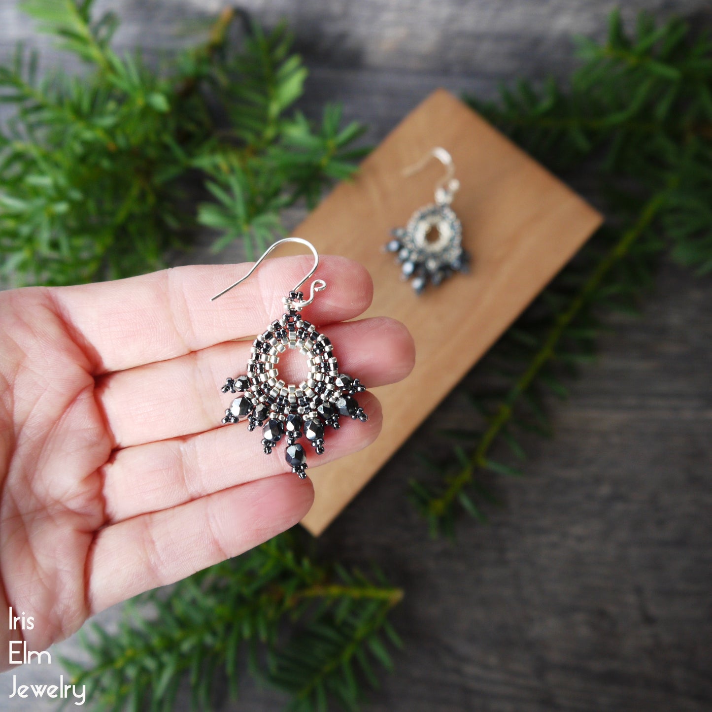 Silver and hematite Czech glass beadwork earrings held in a hand with a wooden block and greenery in the background
