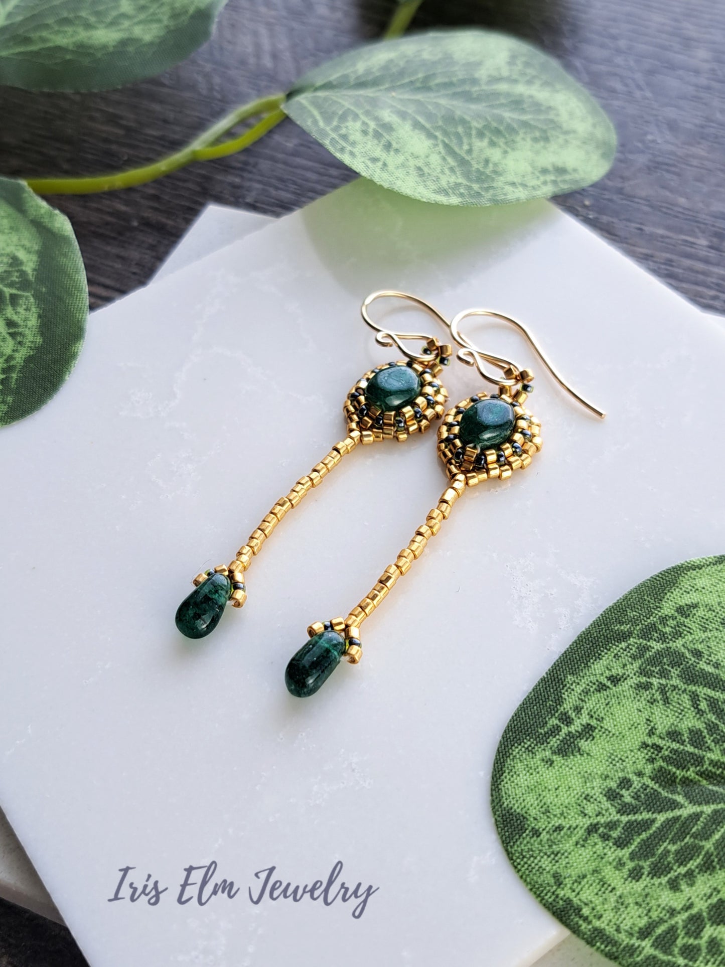 Gold beadwork handmade earrings with green aventurine gemstones on a white napkin with green leaves.