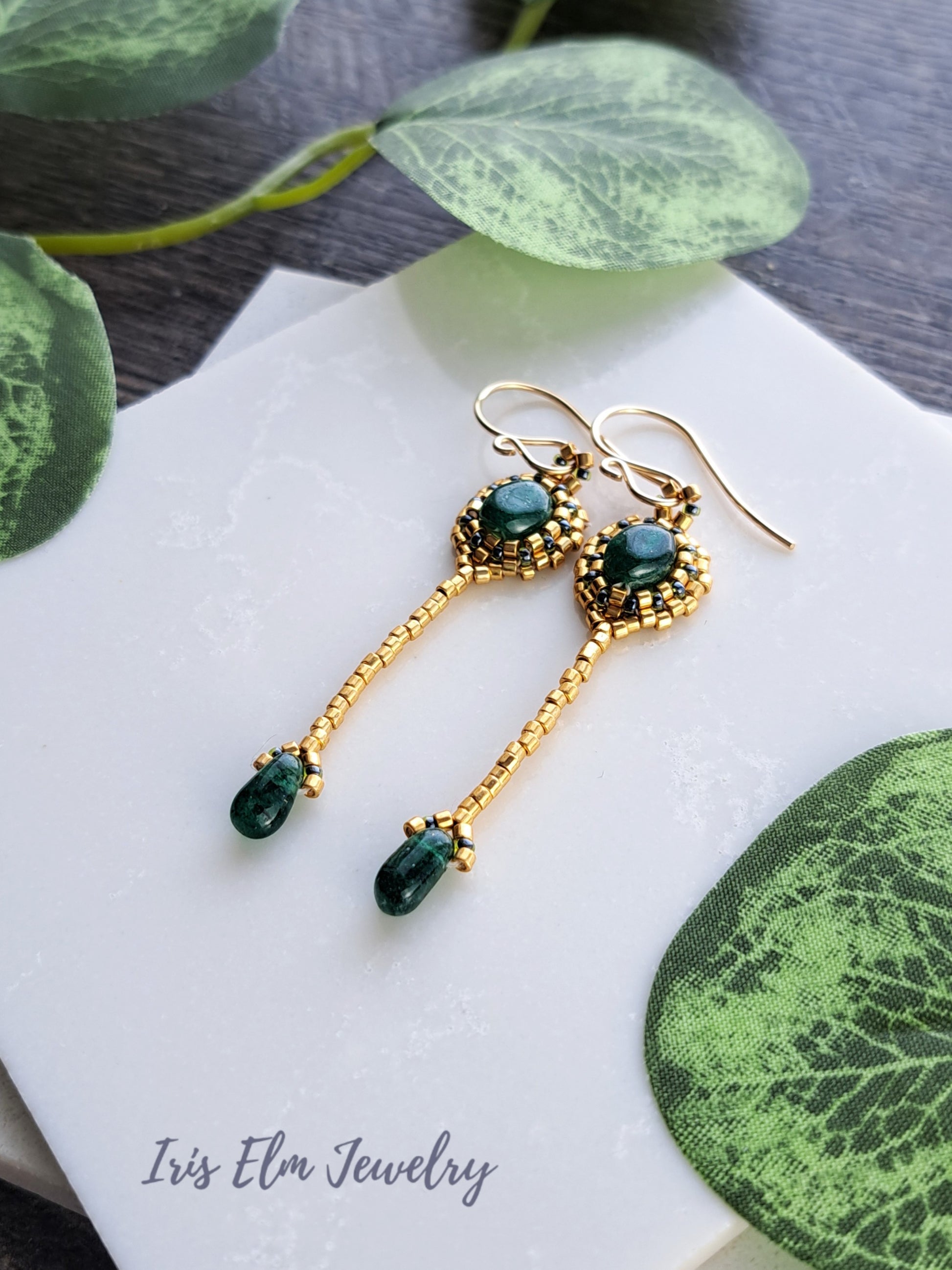 Gold beadwork handmade earrings with green aventurine gemstones on a white napkin with green leaves.
