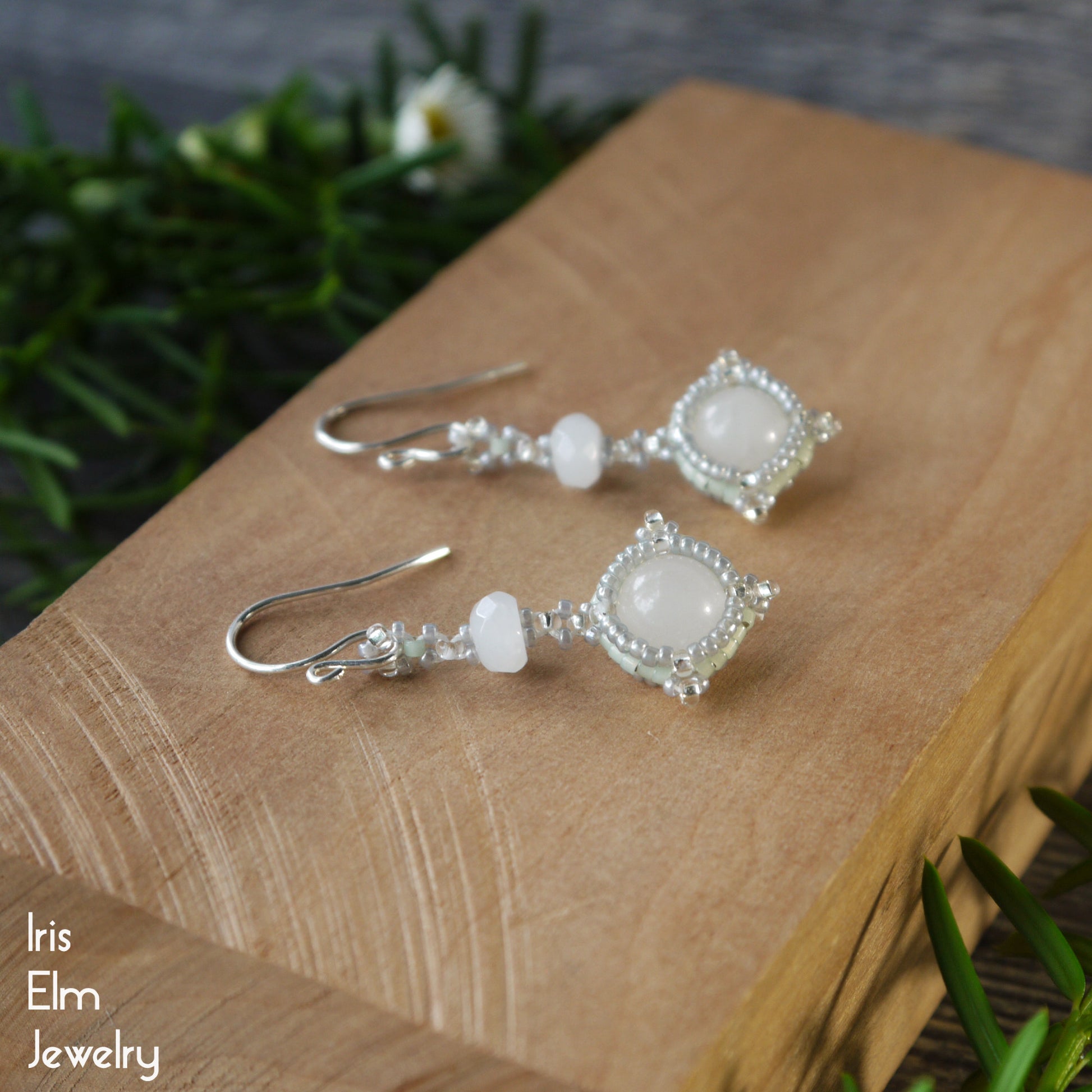 Pair of beadwork white quartz stone and glass seed bead earrings on a wooden surface, branded 'Iris Elm Jewelry'.
