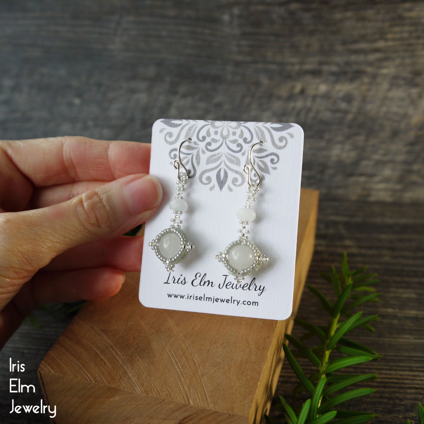Iris Elm Jewelry quartz earrings on a card held by a hand with a wooden surface and plant in the background.