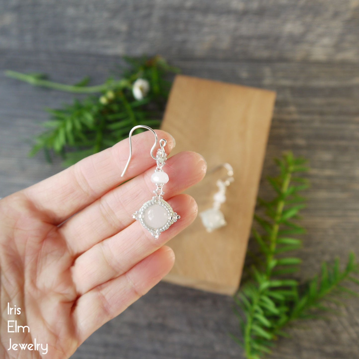 Hand holding a semi-translucent white quartz earring, with a wooden block and greenery in the background.