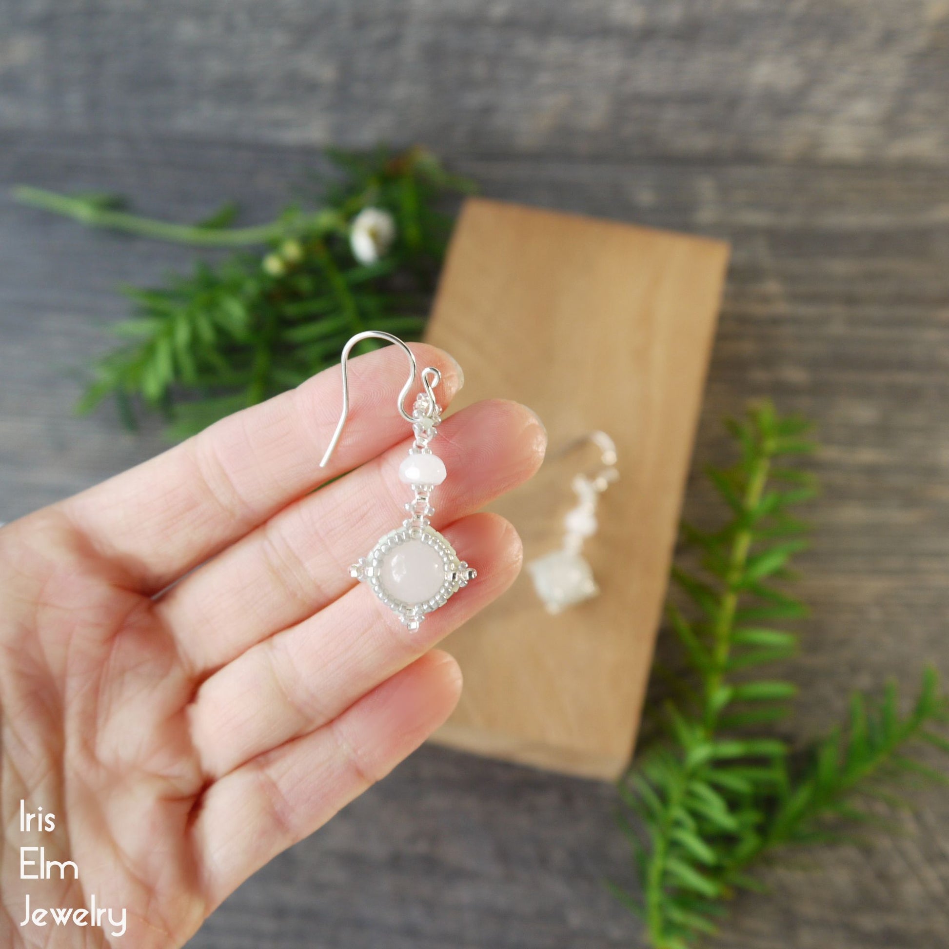 Hand holding a semi-translucent white quartz earring, with a wooden block and greenery in the background.