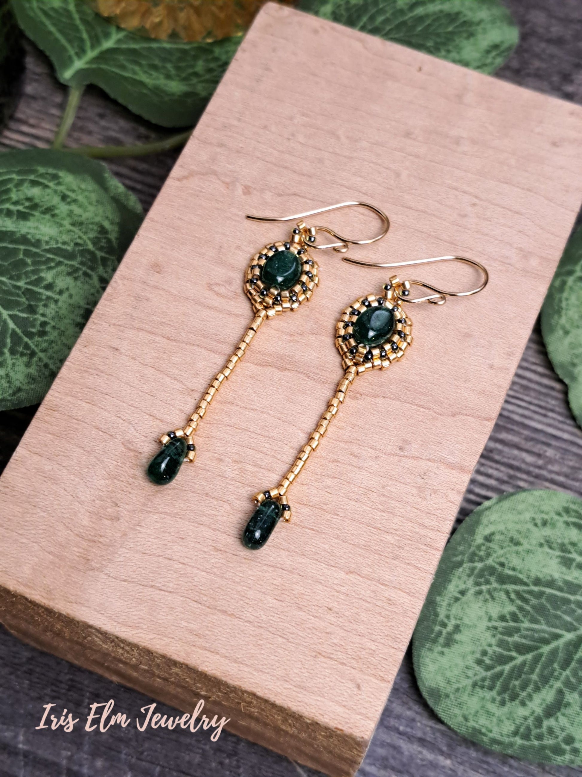 Gold earrings with green aventurine gemstones on a wooden block with leaves in the background