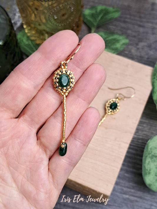 Gold earrings with green aventurine gemstones held in a hand, brand 'Iris Elm Jewelry' visible.