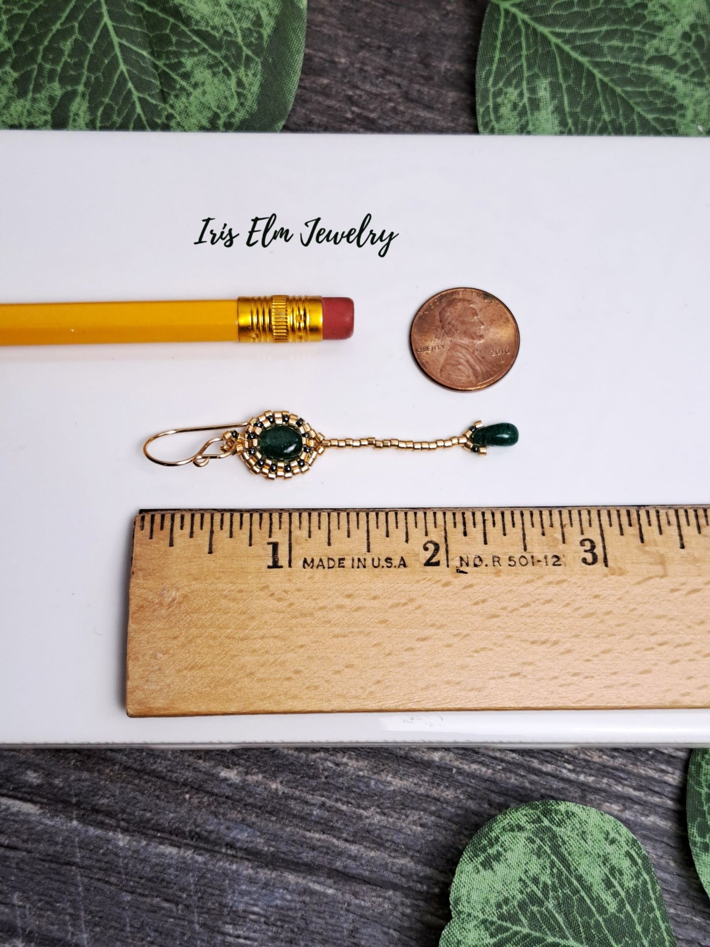 2.5 inch long earrings with green gemstone next to a pencil and coin for scale, on a wooden surface with 'Iris Elm Jewelry' branding.