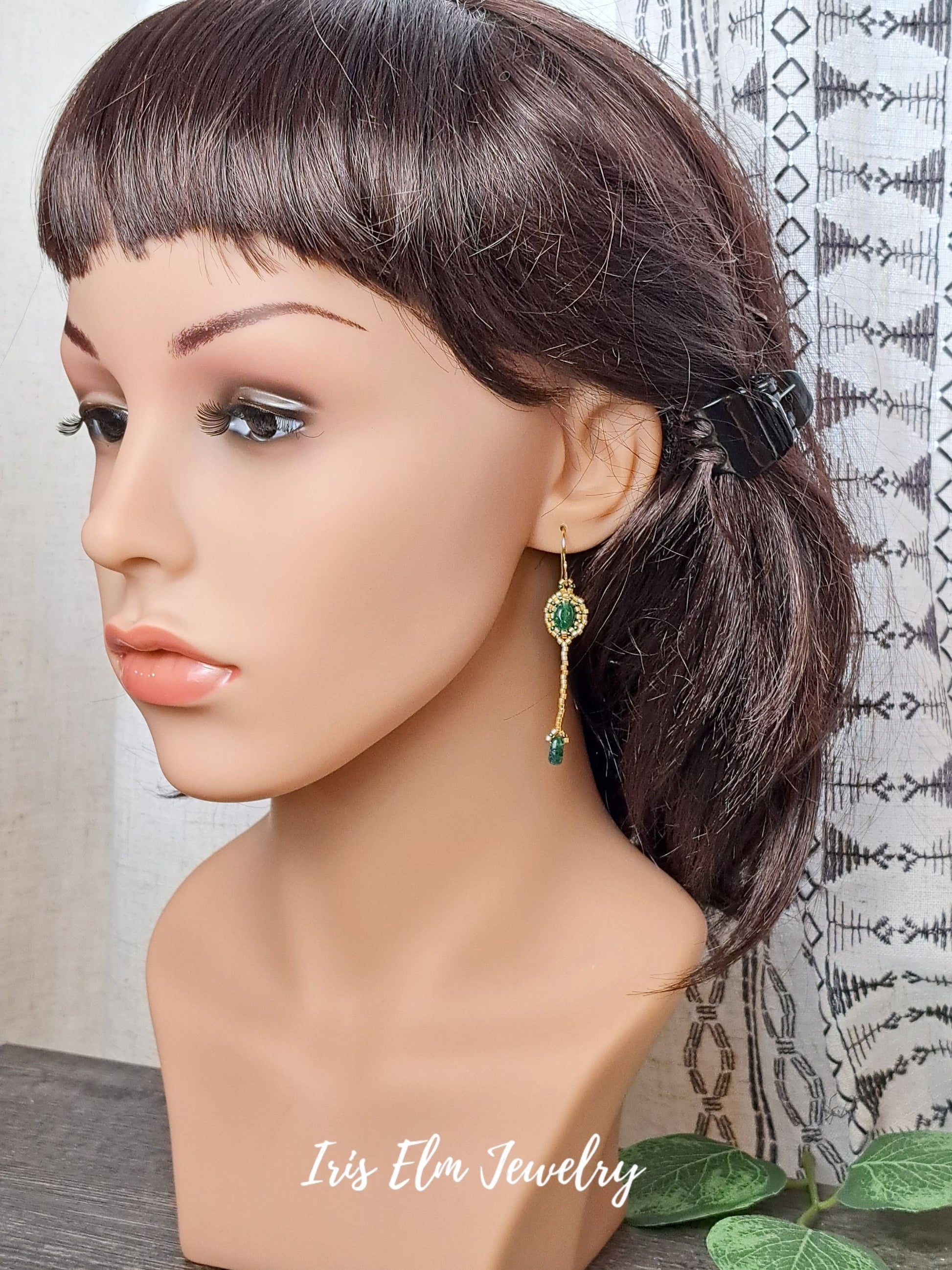 Mannequin head with dark hair styled in a ponytail, wearing a green aventurine earring, with 'Iris Elm Jewelry' branding.