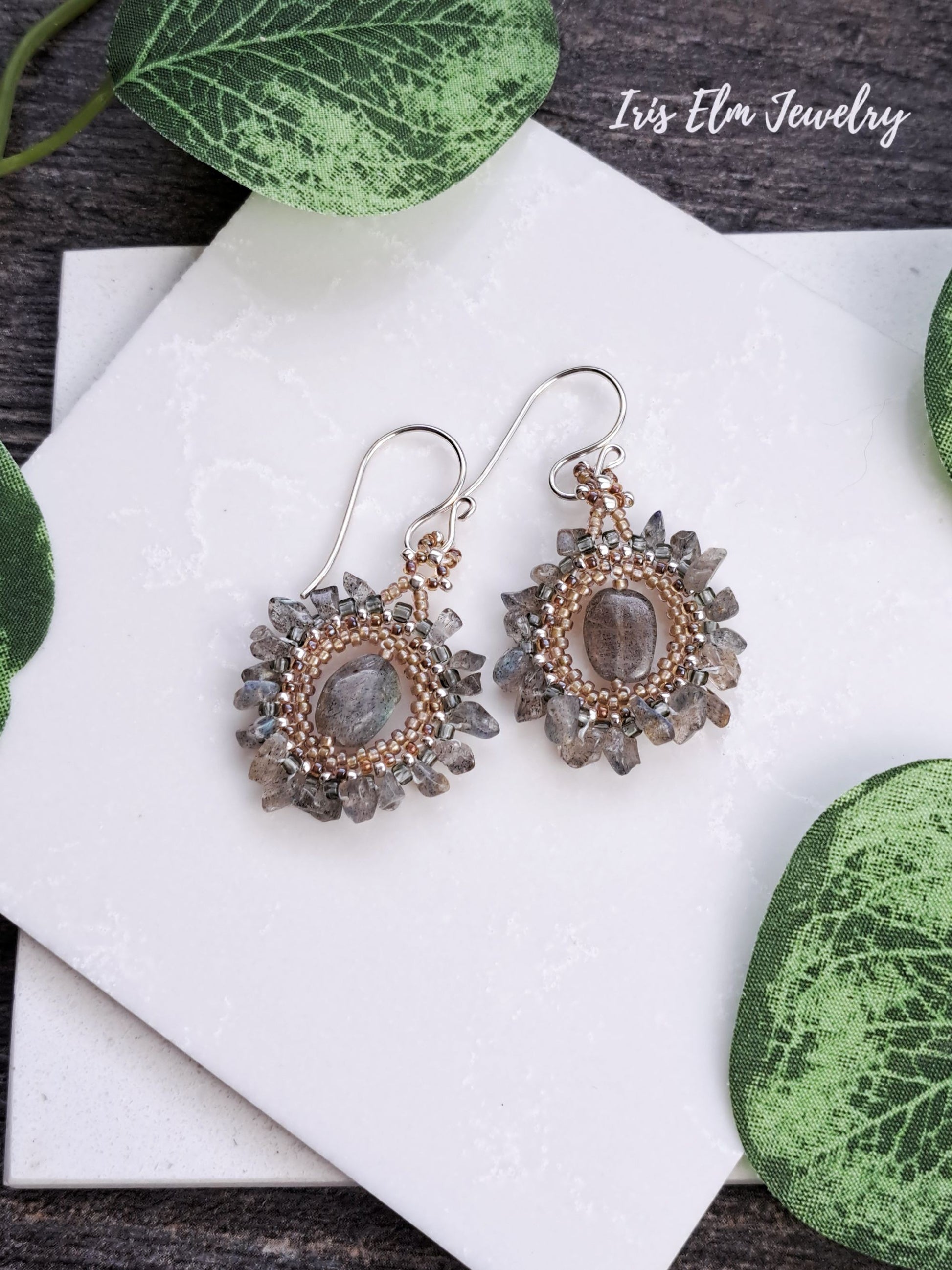 Pair of bohemian labradorite beadwork handmade earrings with gray stones on a white surface with green leaves.