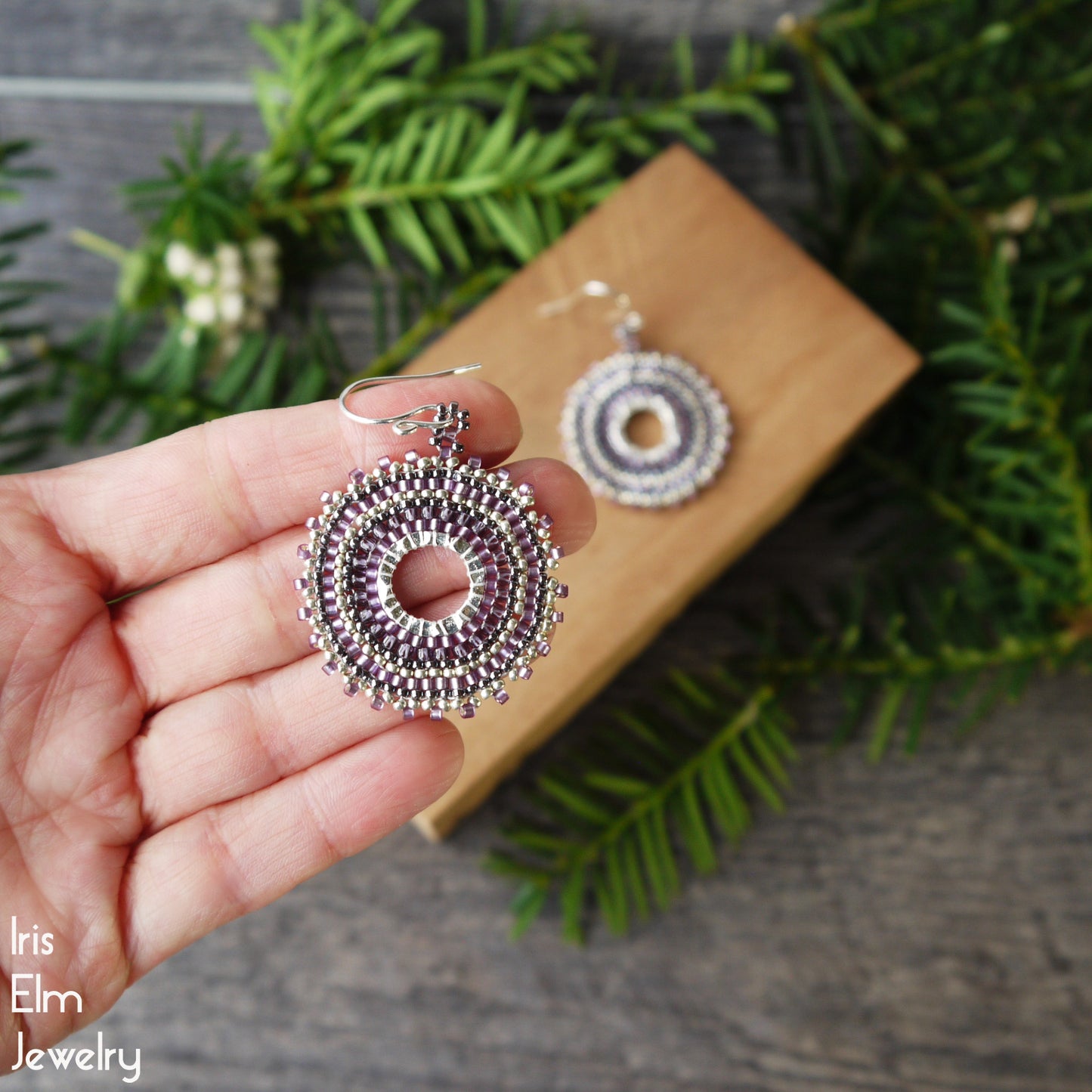 Hand holding a silver and purple beaded circular earring with a wooden box and greenery in the background.