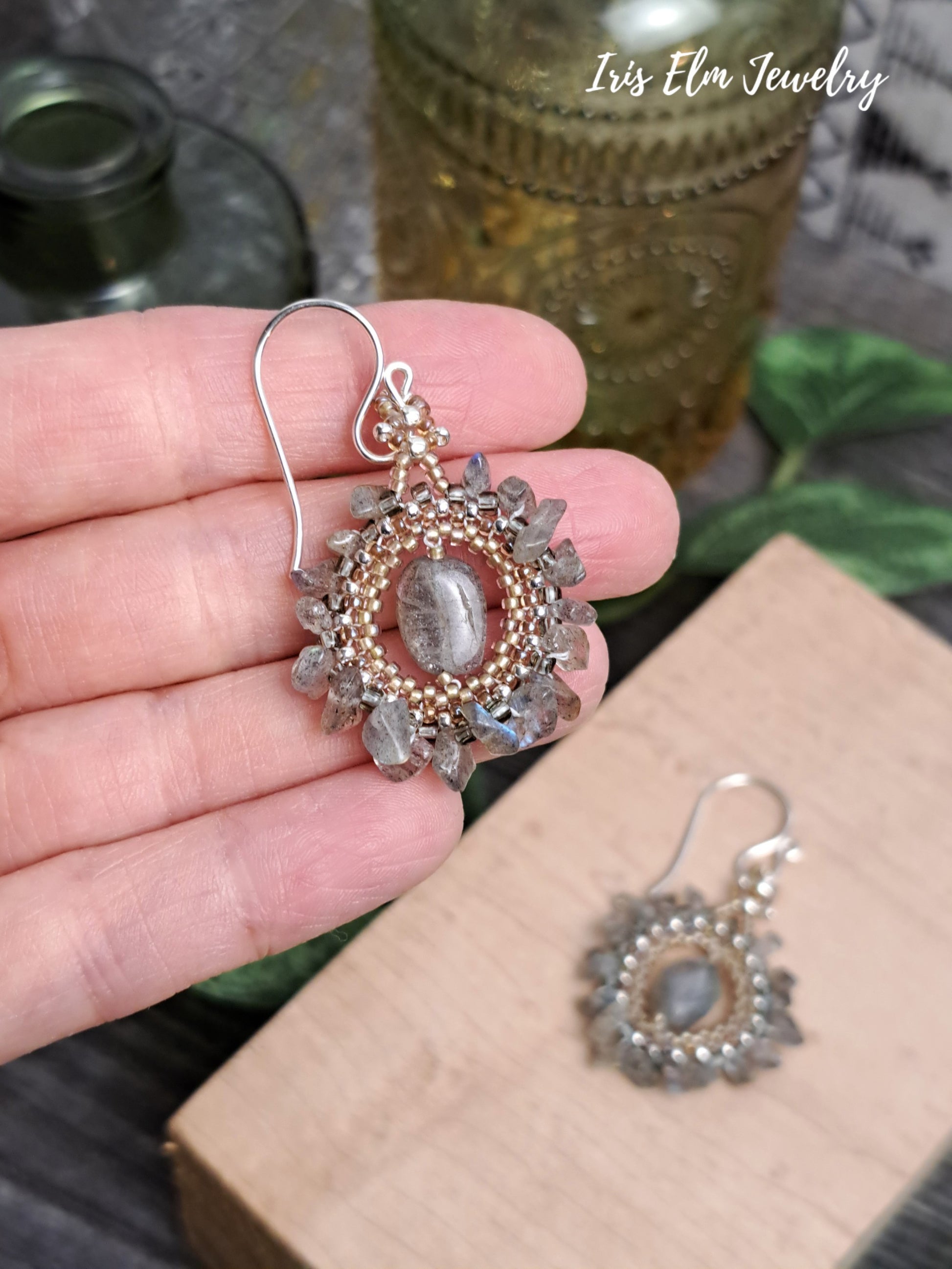 Hand holding a pair of bohemian labradorite beadwork handmade earrings with gemstones, branded 'Iris Elm Jewelry', on a rustic background.