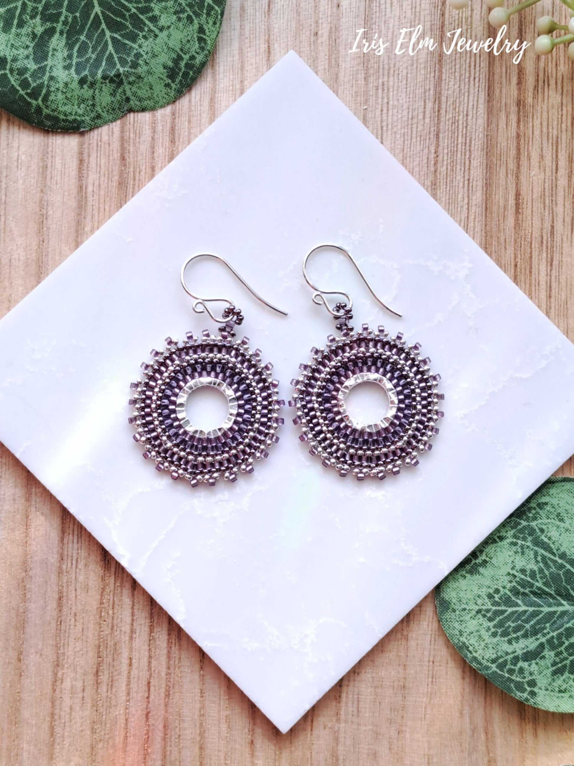 Pair of silver and purple woven seed bead hoop earrings with purple beads on a white card with green leaves around