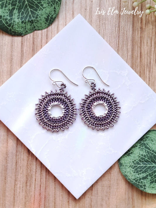 Pair of silver and purple woven seed bead hoop earrings with purple beads on a white card with green leaves around