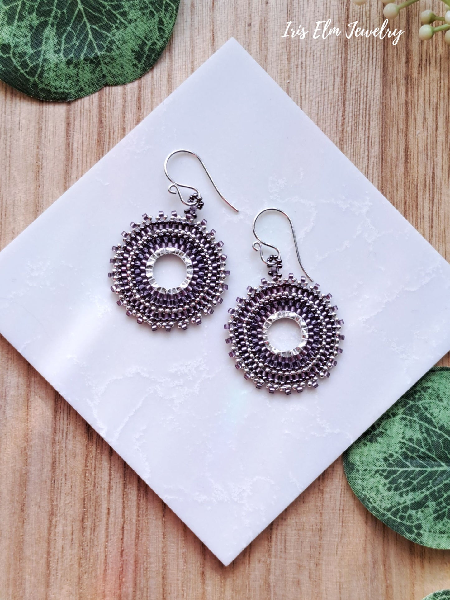 Decorative purple woven seed bead hoop earrings on a white card with green leaves in the background