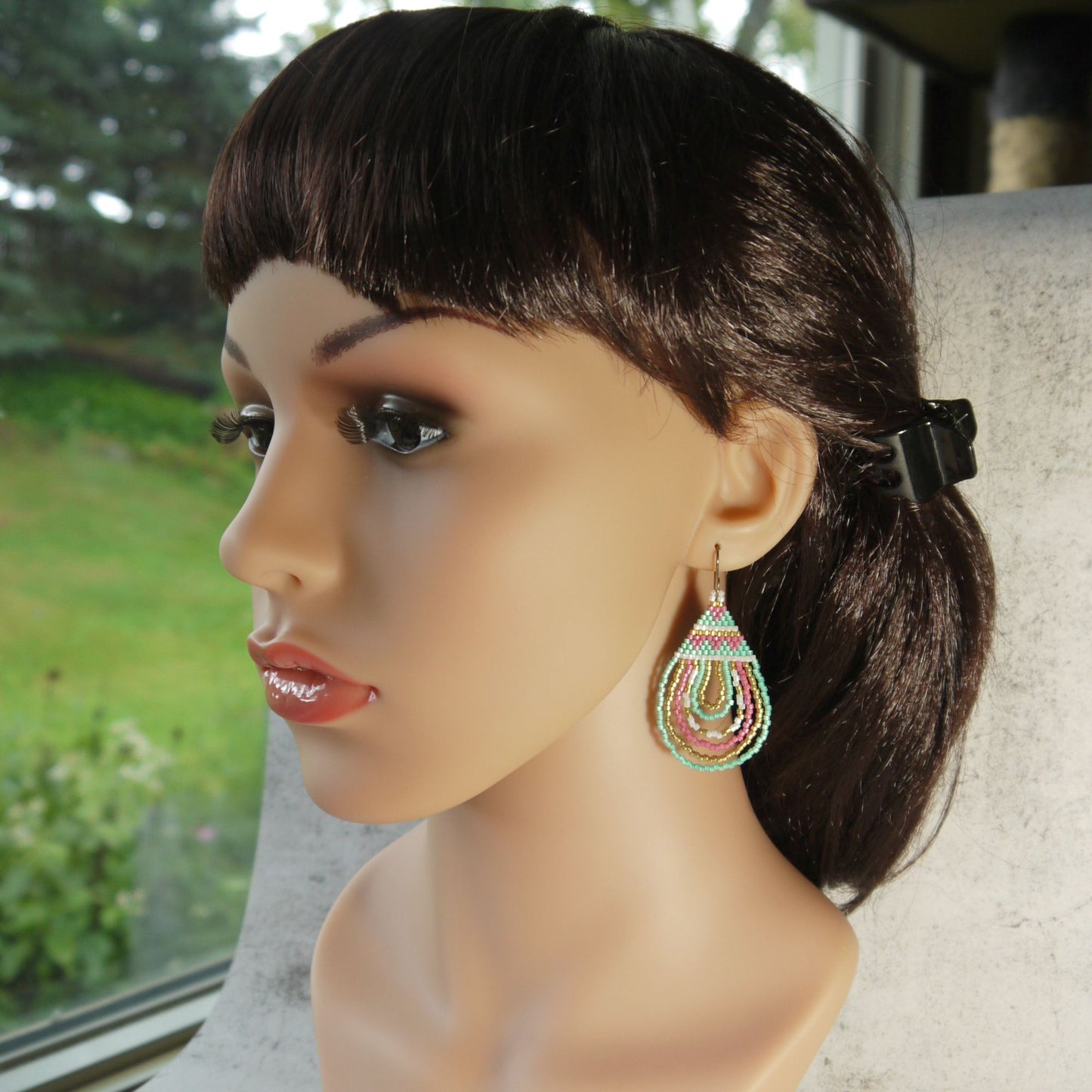 Mannequin head with dark hair and colorful pink and green earrings, blurred greenery in the background