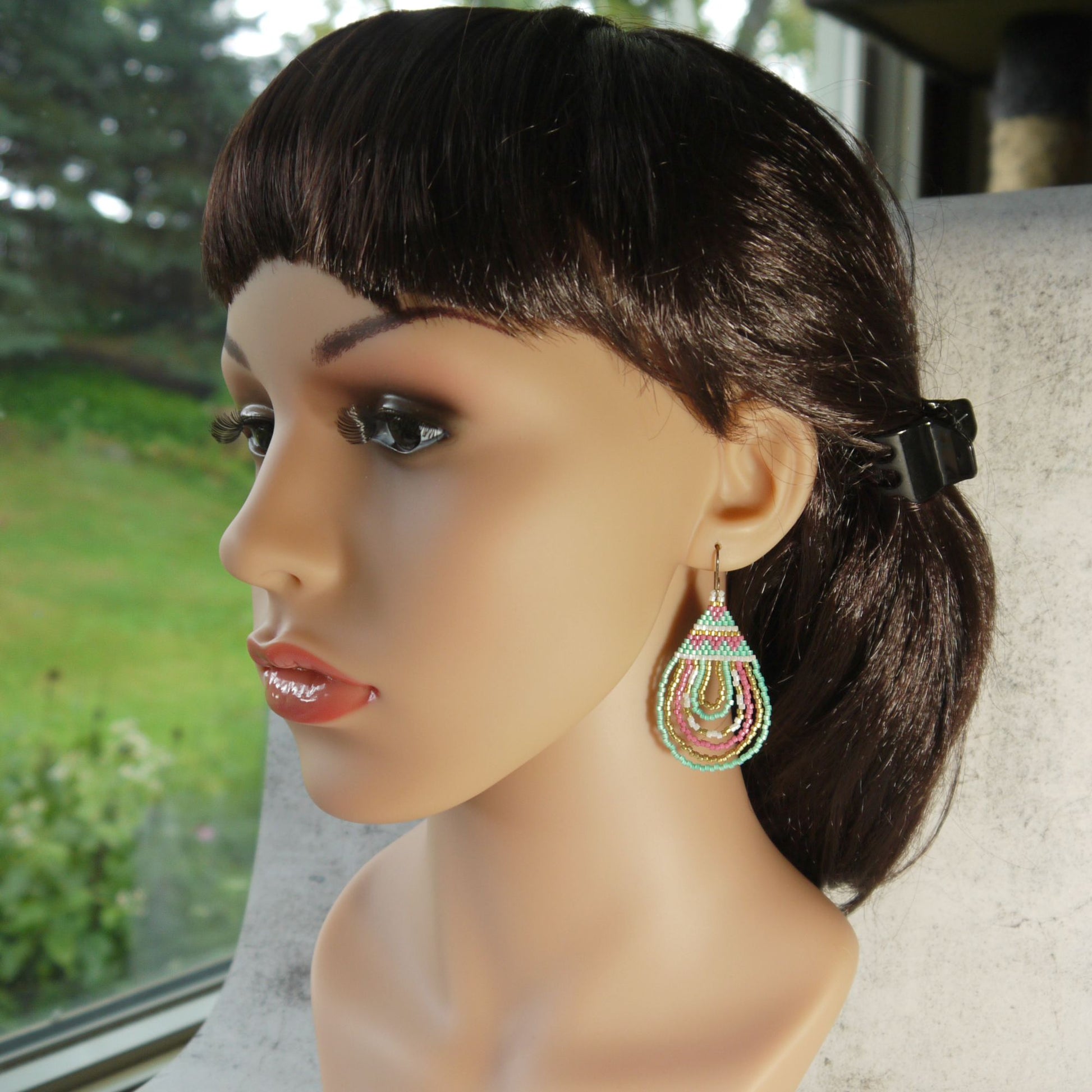 Mannequin head with dark hair and colorful pink and green earrings, blurred greenery in the background