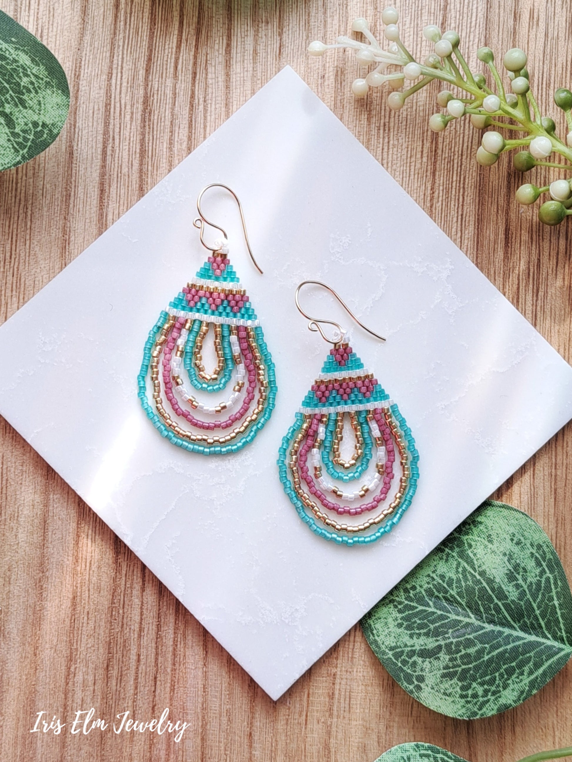 Colorful beaded mint green, pink, and gold teardrop seed bead earrings on a white card with green leaves and flowers in the background