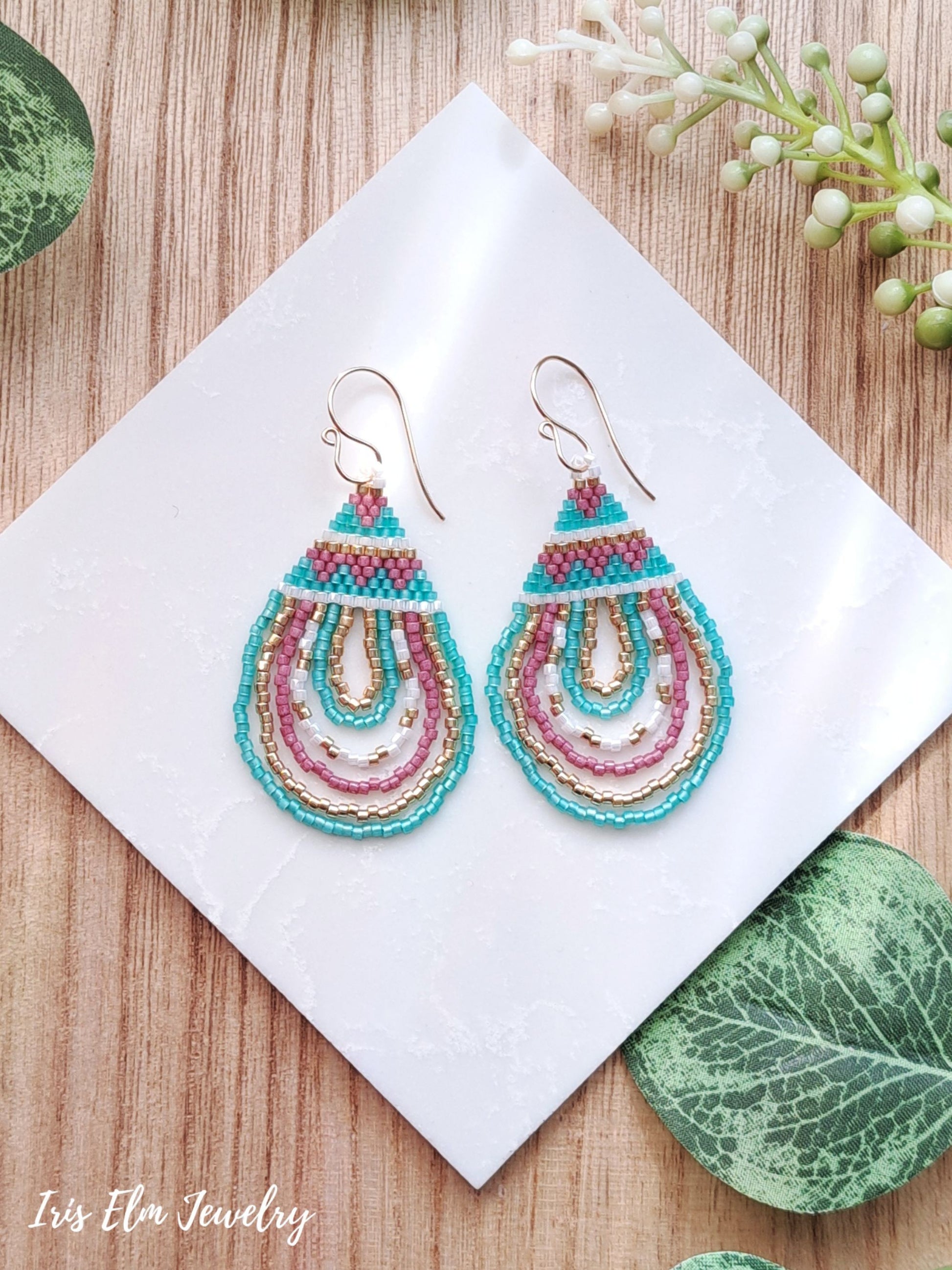 Colorful beaded pink, mint, and gold seed bead teardrop earrings on a white card with green leaves and flowers in the background