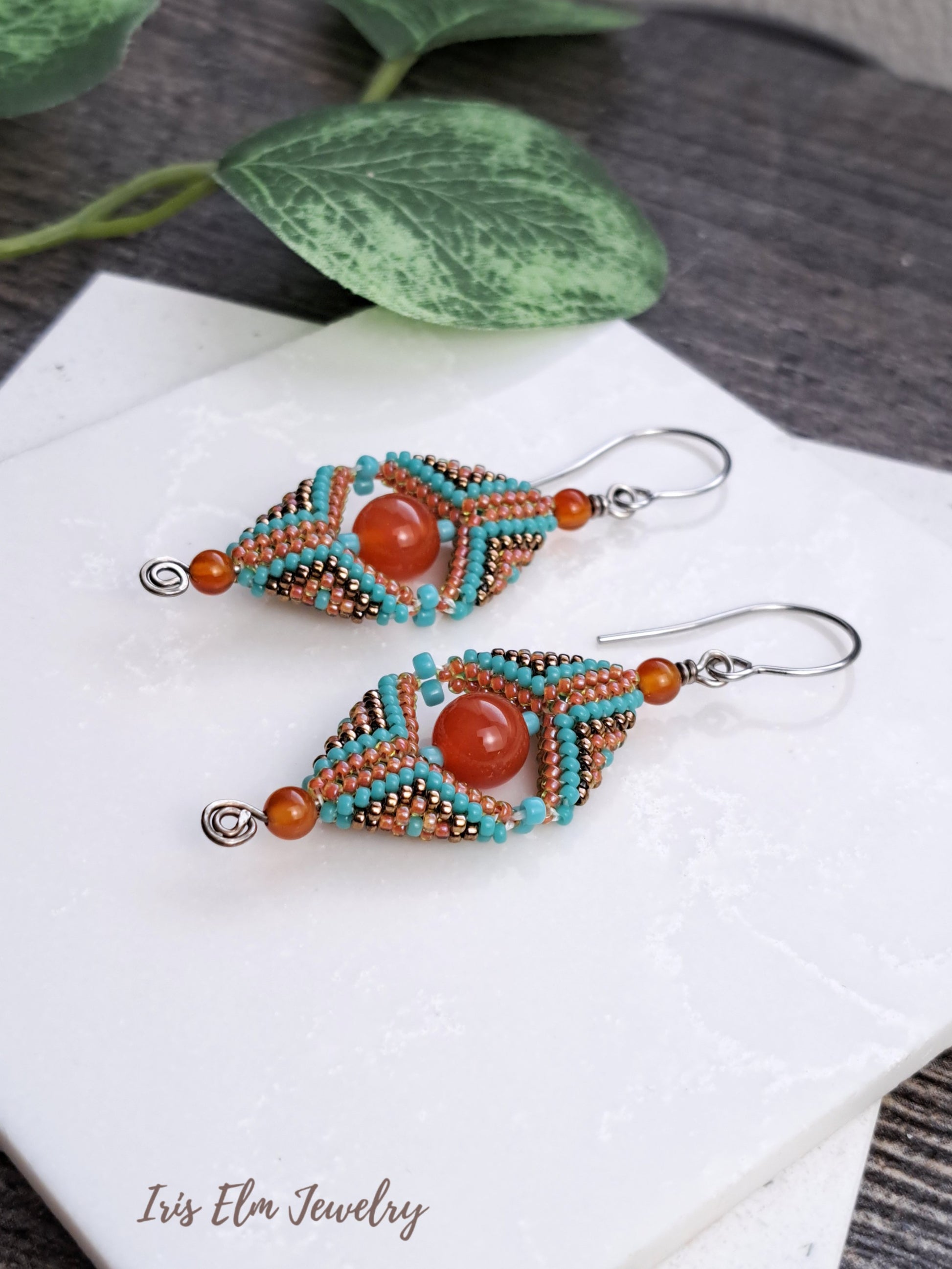 Beaded carnelian gemstone and seed bead beadwork earrings with turquoise and orange beads on a white surface with green leaves in the background.