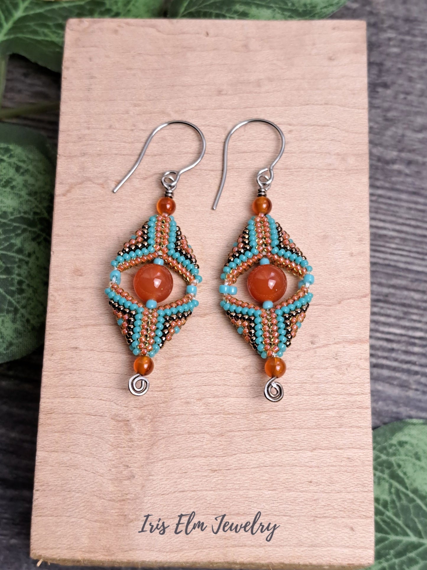 Beaded handmade carnelian gemstone and seed bead beadwork earrings on a wooden card with 'Iris Elm Jewelry' branding, surrounded by green leaves.