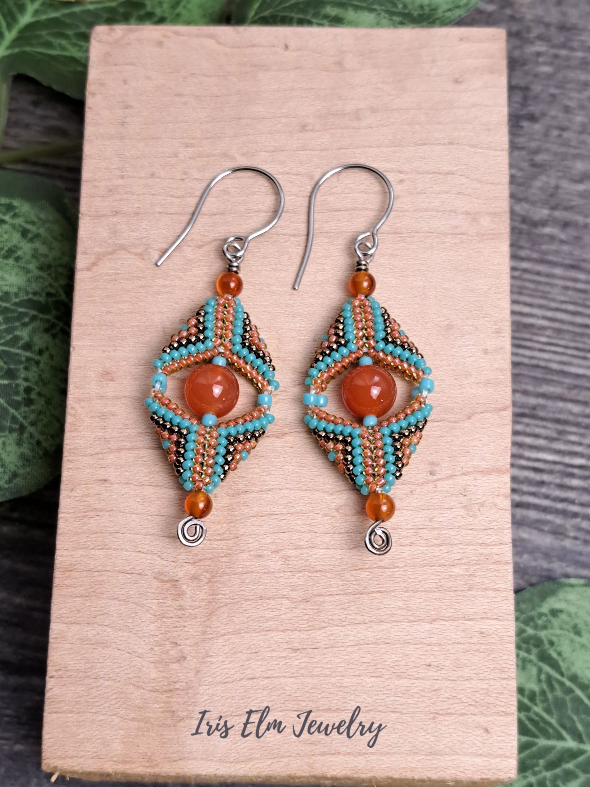 Beaded handmade carnelian gemstone and seed bead beadwork earrings on a wooden card with 'Iris Elm Jewelry' branding, surrounded by green leaves.