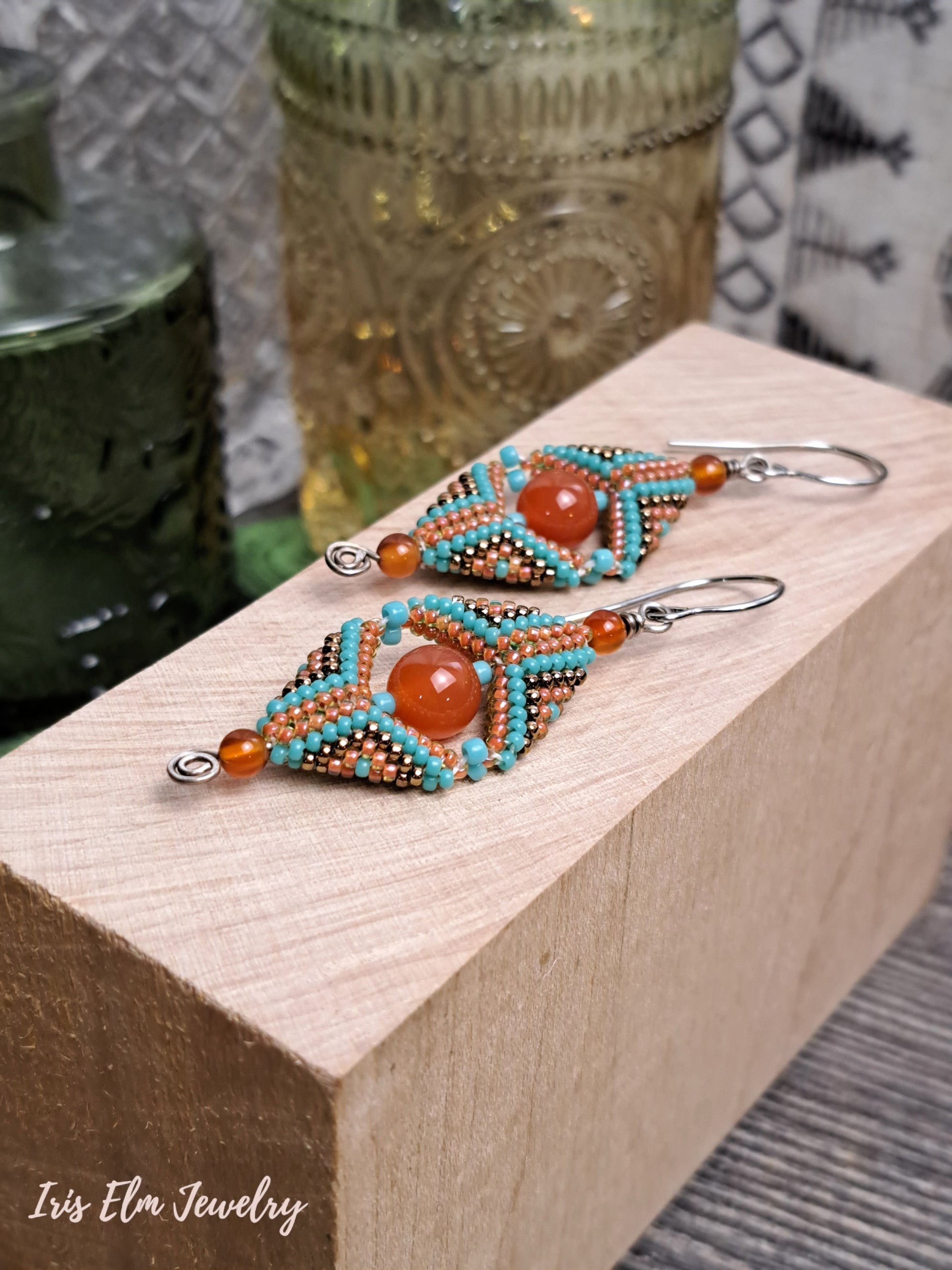 Beaded carnelian gemstone and seed bead beadwork earrings on a wooden block with 'Iris Elm Jewelry' branding.