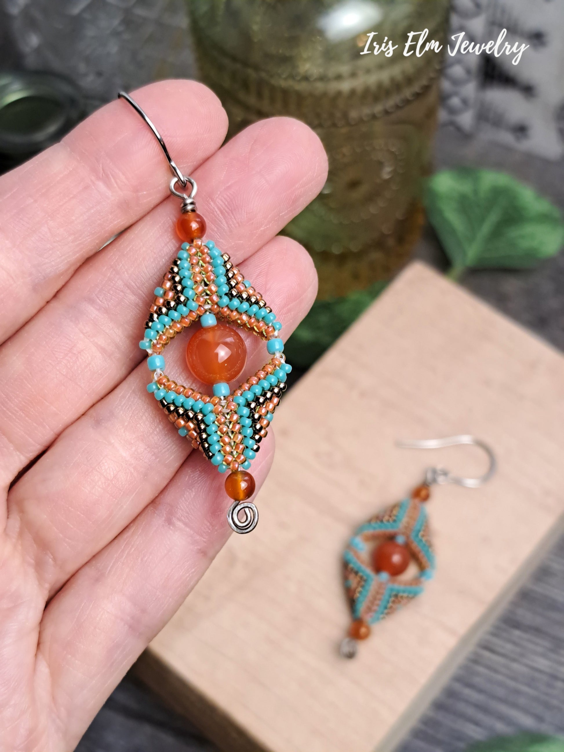Beaded carnelian gemstone and seed bead beadwork earrings with orange and turquoise design held by a hand, with 'Iris Elm Jewelry' branding.