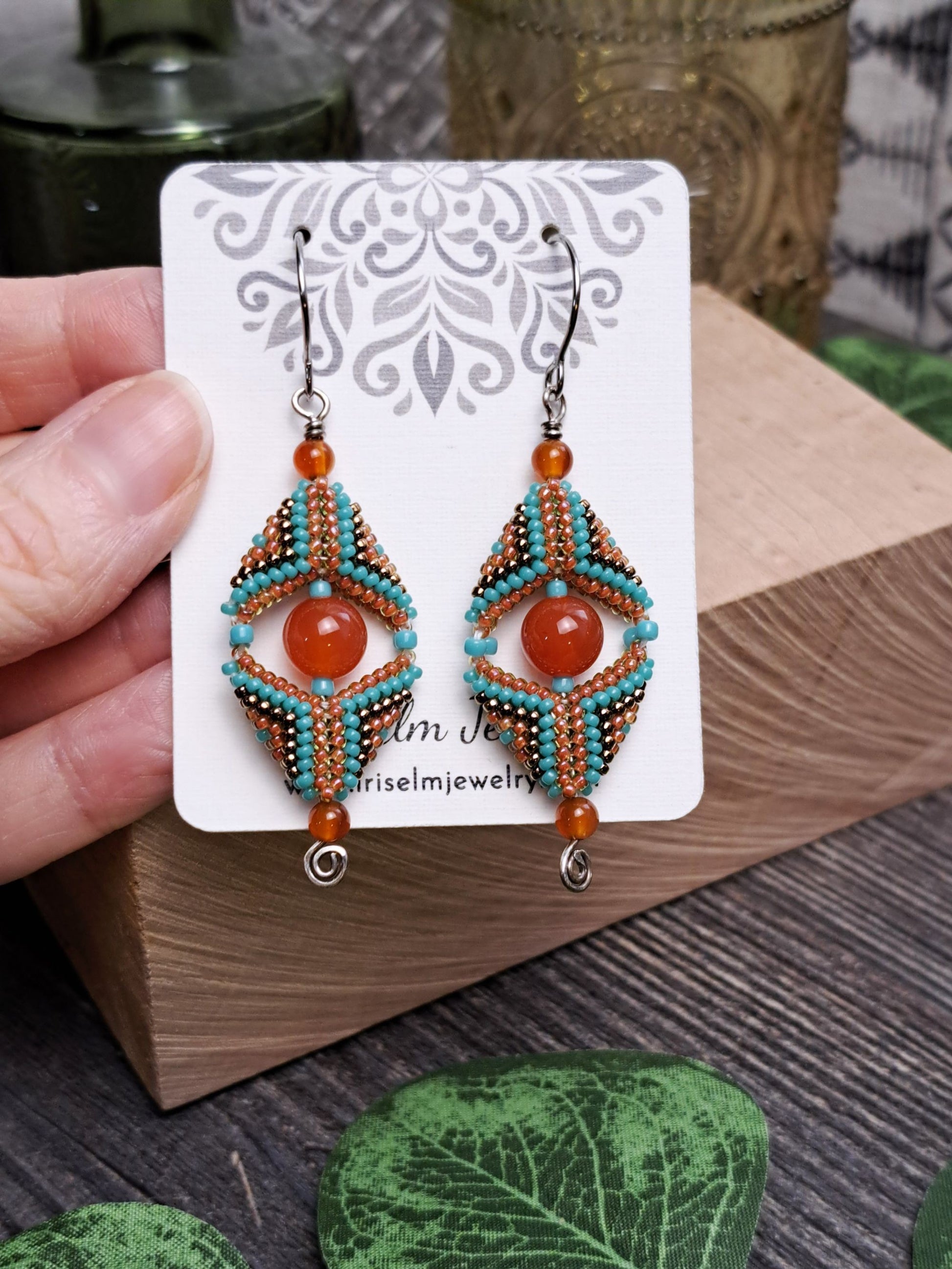 Pair of carnelian gemstone and seed bead beadwork beaded earrings on a decorative card held by a hand.