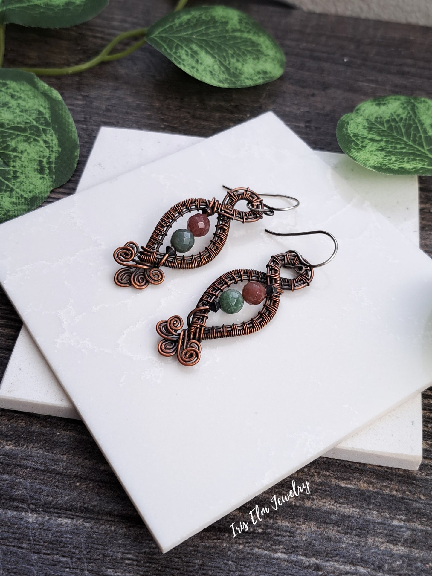 Pair of copper wire-wrapped earrings with green stones on a white marble surface.
