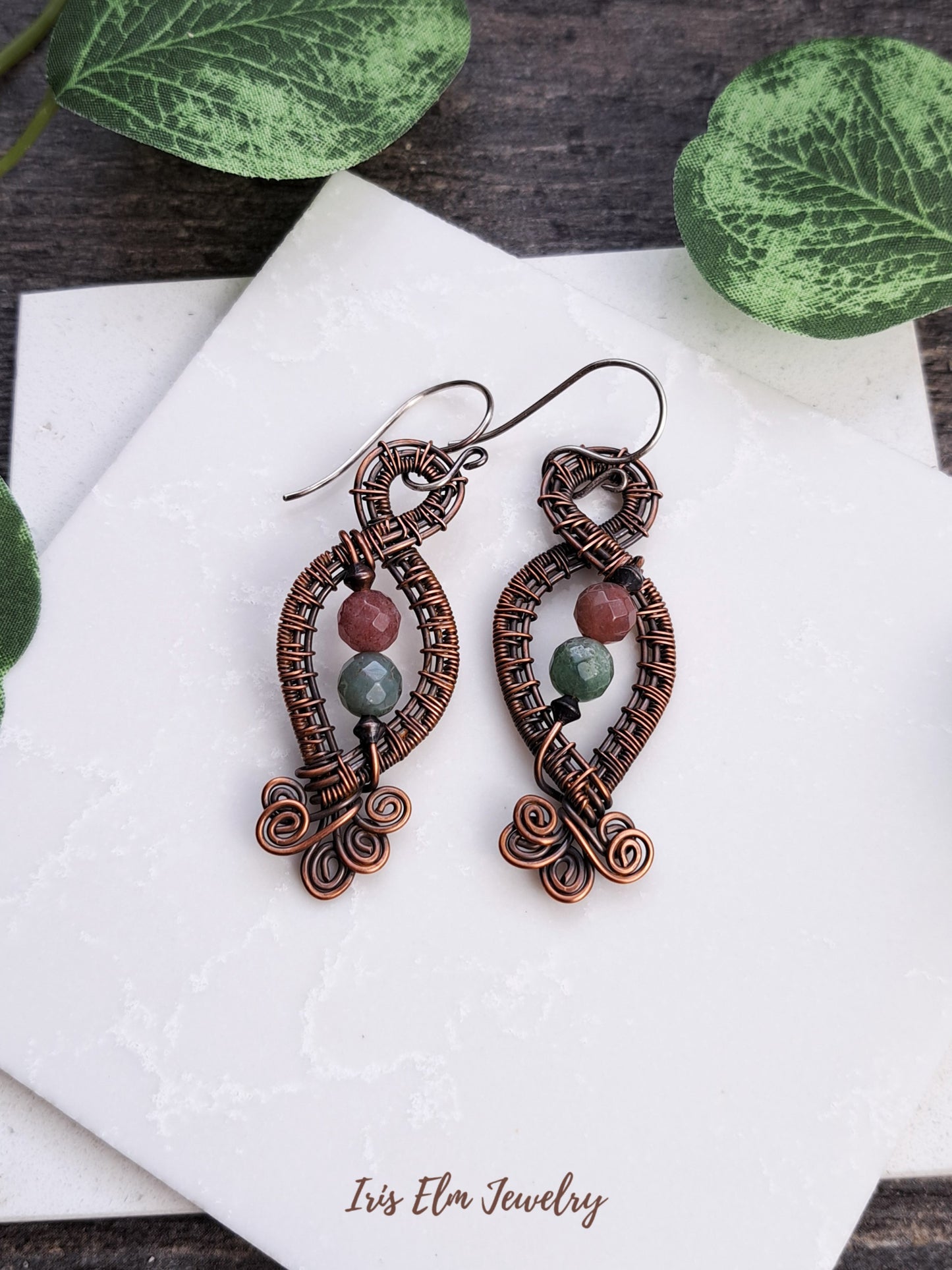 Pair of intricate handmade copper earrings with green and mauve jasper beads on a white surface with leaves in the background.