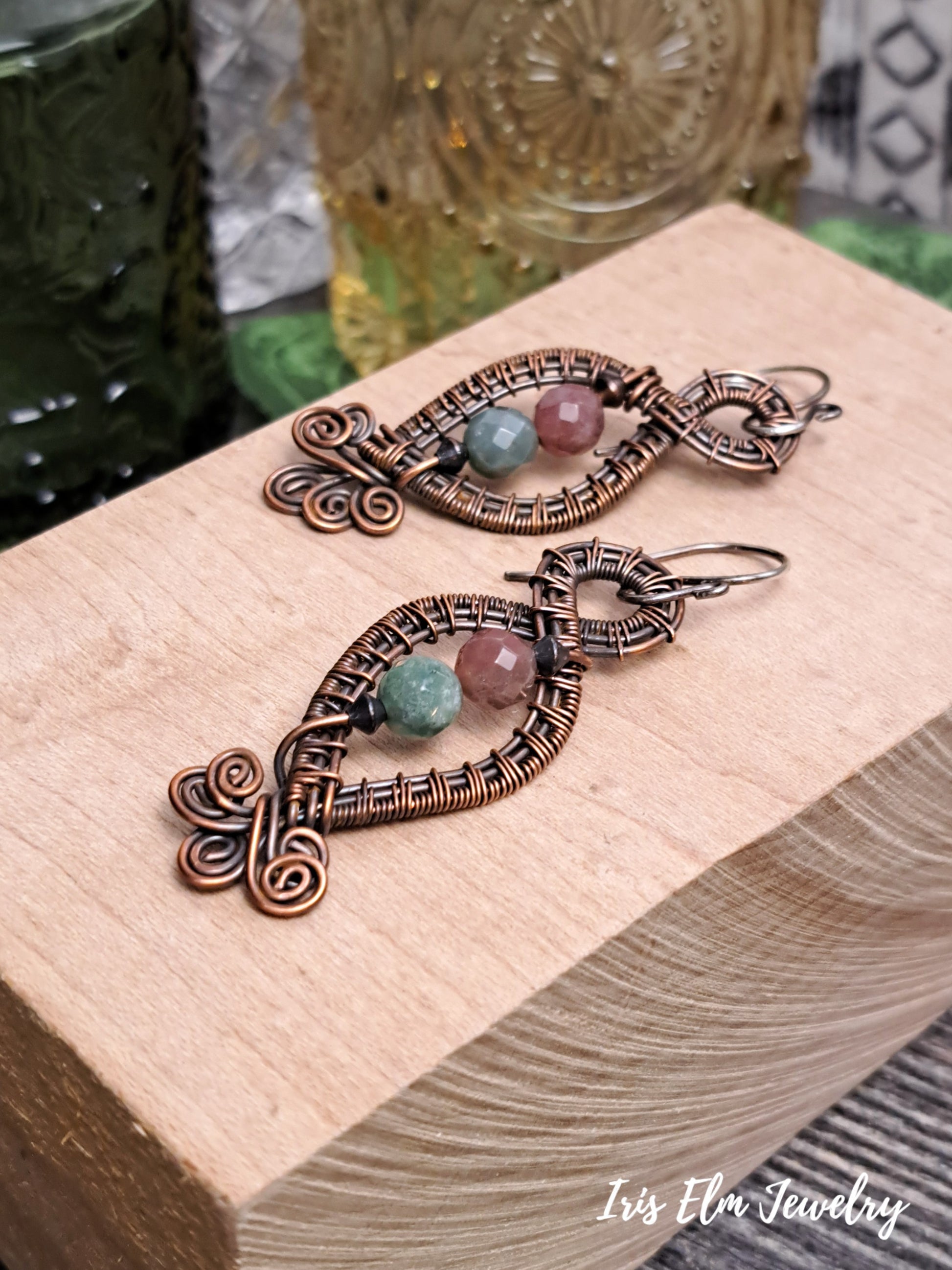 Pair of copper earrings with jasper gemstones on a wooden block, brand 'Iris Elm Jewelry' visible.