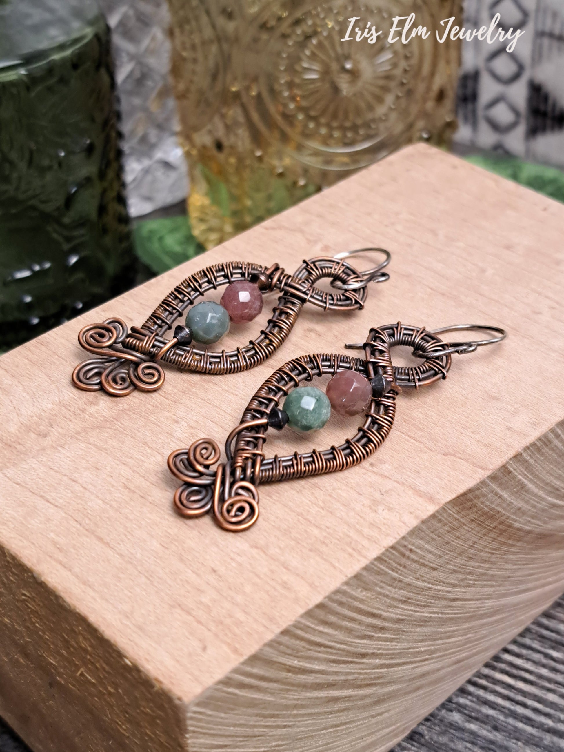 Copper earrings with mauve and green jasper gemstones on a wooden block, branded 'Iris Elm Jewelry'.