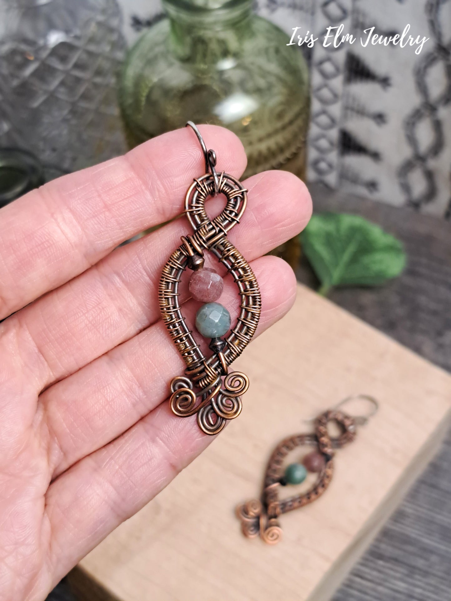 Decorative fancy jasper copper wire woven earrings held in a hand with 'Iris Elm Jewelry' branding.