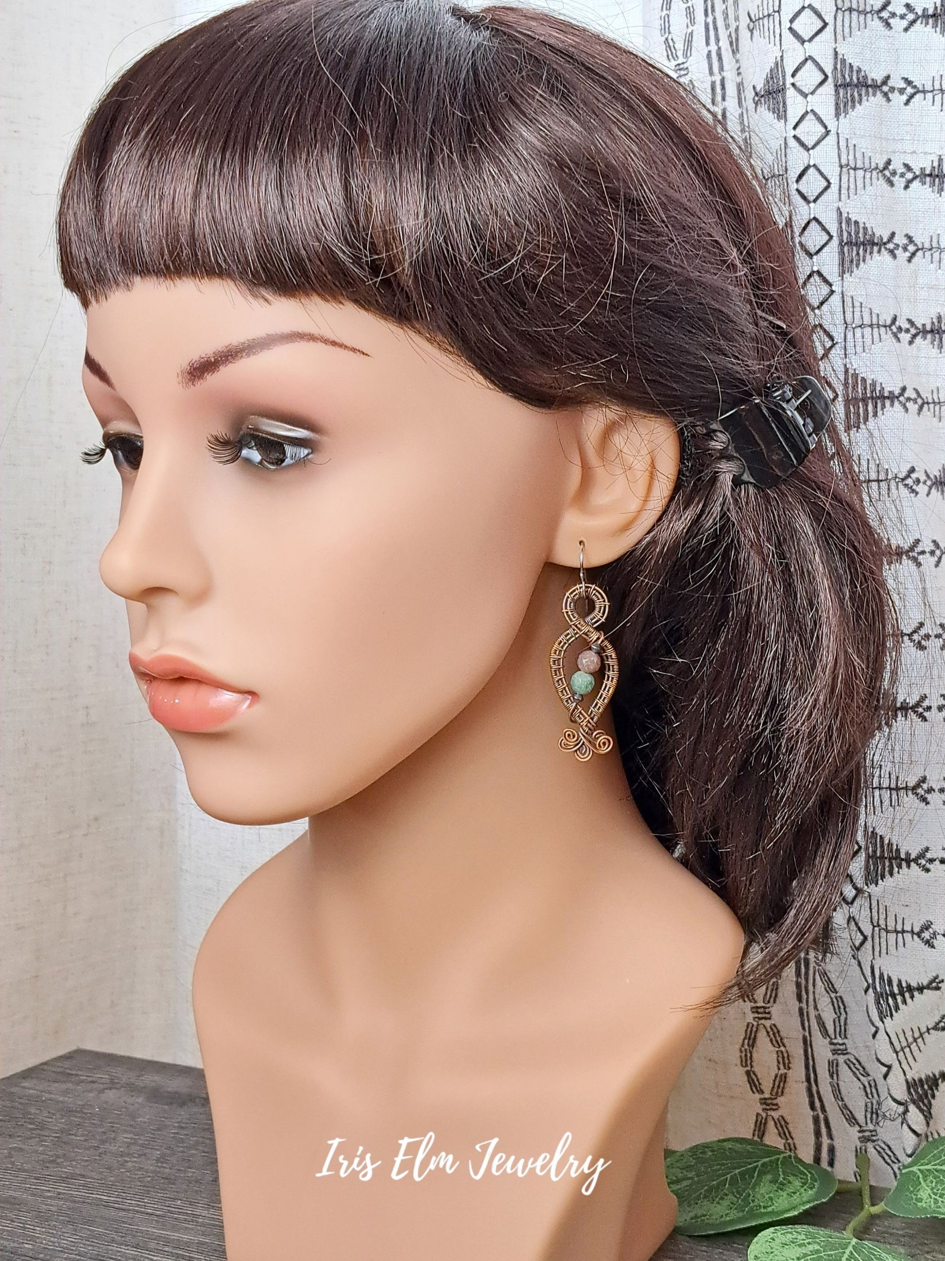 Mannequin head with brown hair styled in a ponytail, wearing jasper copper wire earrings with stones, against a neutral background.