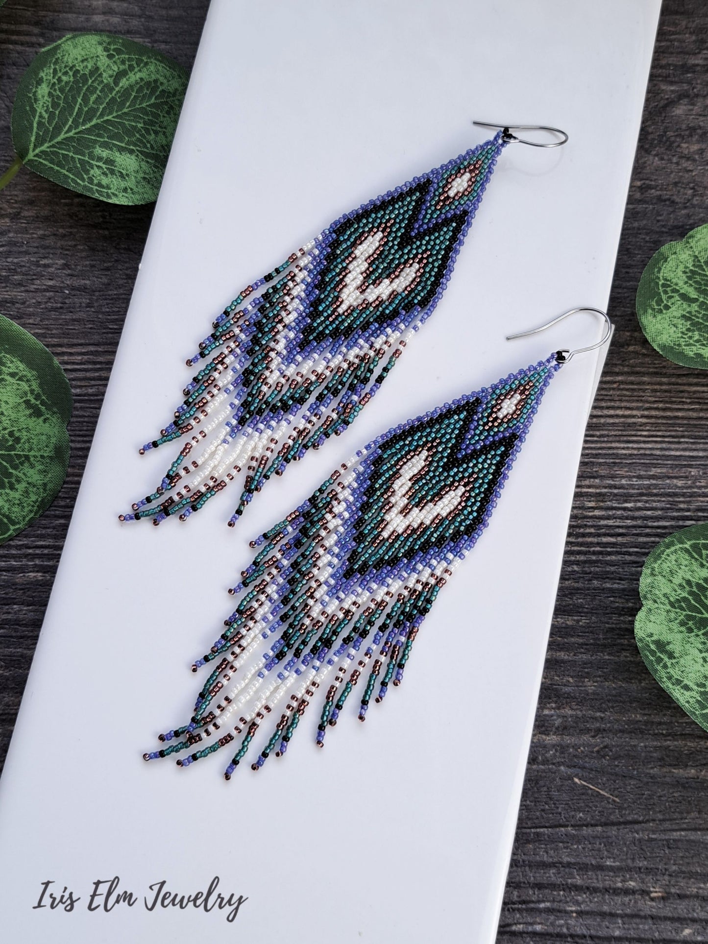 long purple, teal, black, and white seed bead heart fringe Beaded earrings on a white tile with green leaves in the background