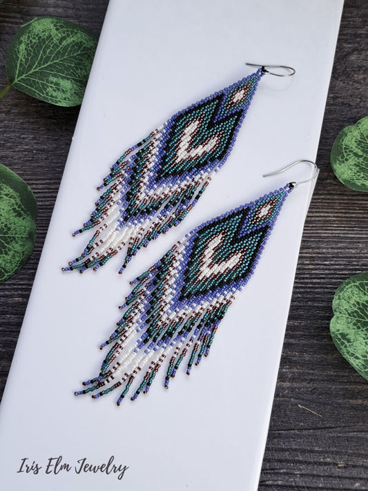 long purple, teal, black, and white seed bead heart fringe Beaded earrings on a white tile with green leaves in the background