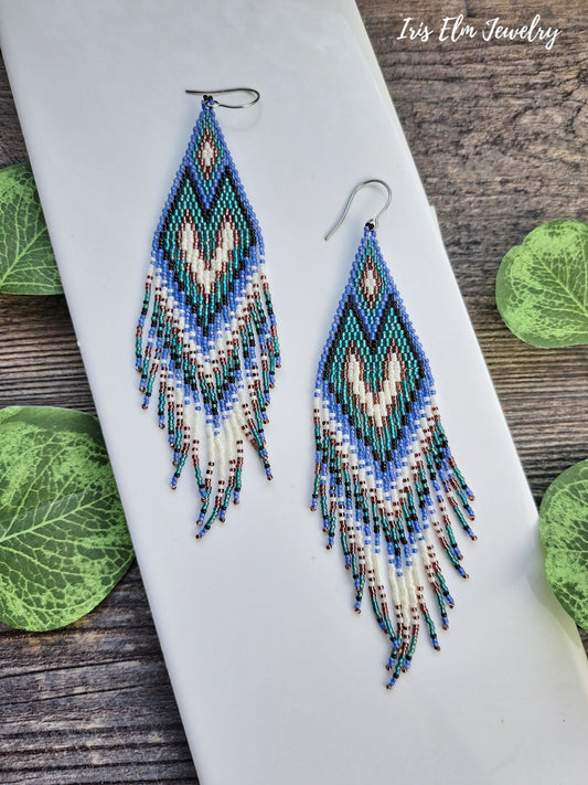 Beaded long purple, teal, black, and white seed bead heart fringe earrings on a white surface with green leaves in the background, featuring the brand 'Iris Elm Jewelry'.